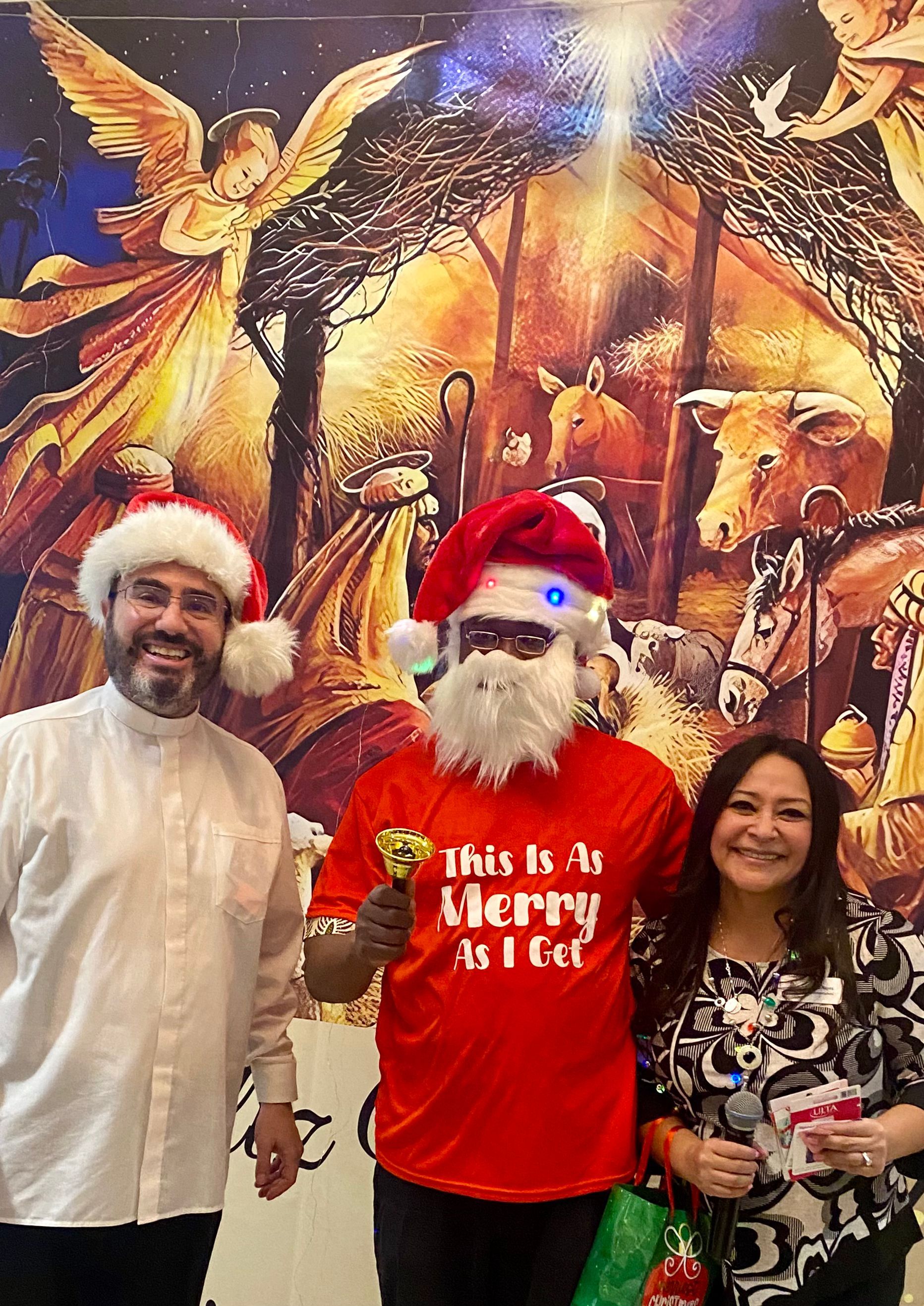 Three people in Santa hats pose before a festive holiday mural; center person wears a red “This is a Merry list” shirt.