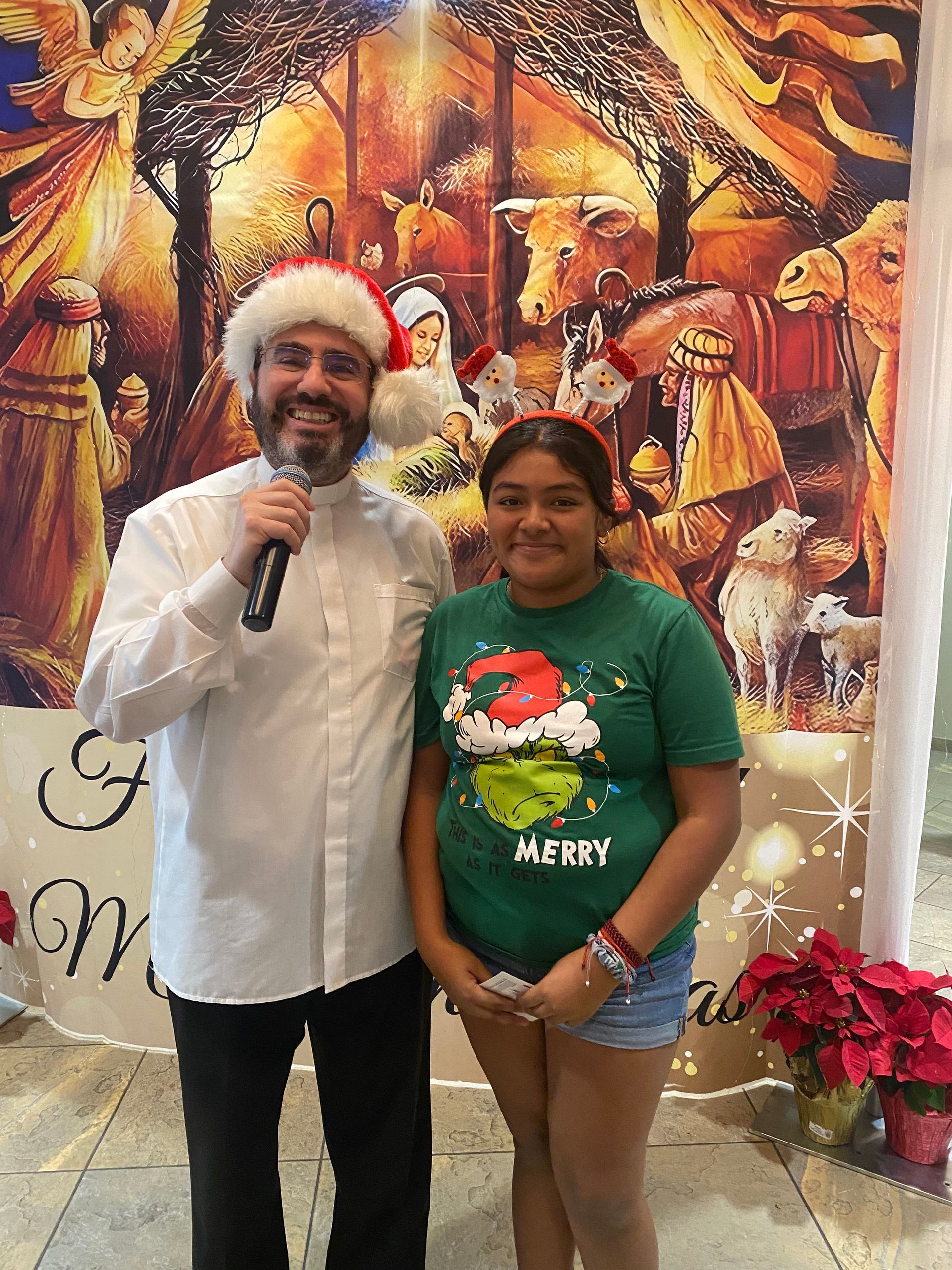 Two people pose smiling at an indoor festive event, with one holding a microphone in front of a holiday backdrop.
