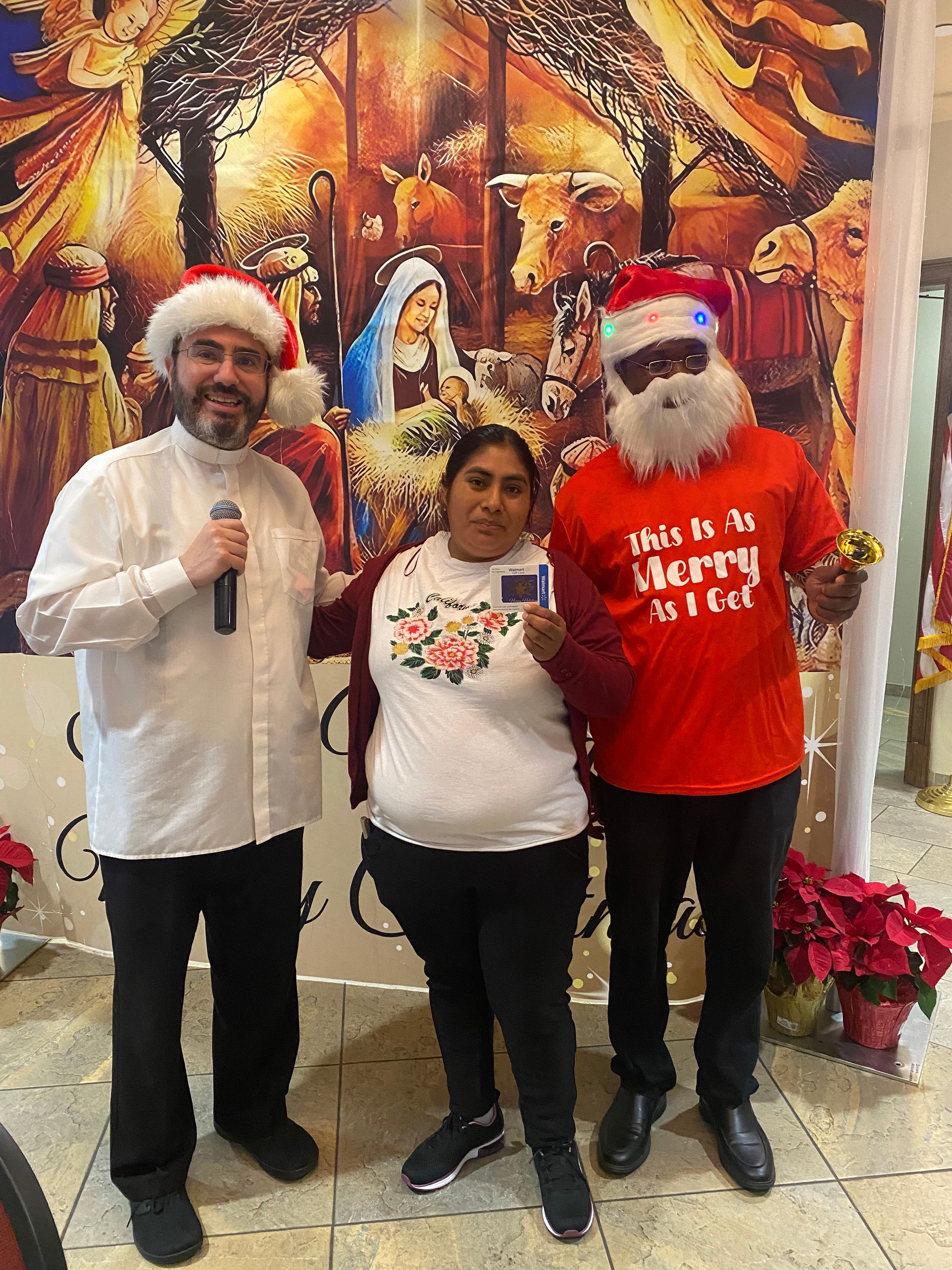 Three people posing indoors in front of a festive mural, two wearing Santa hats and holiday shirts.