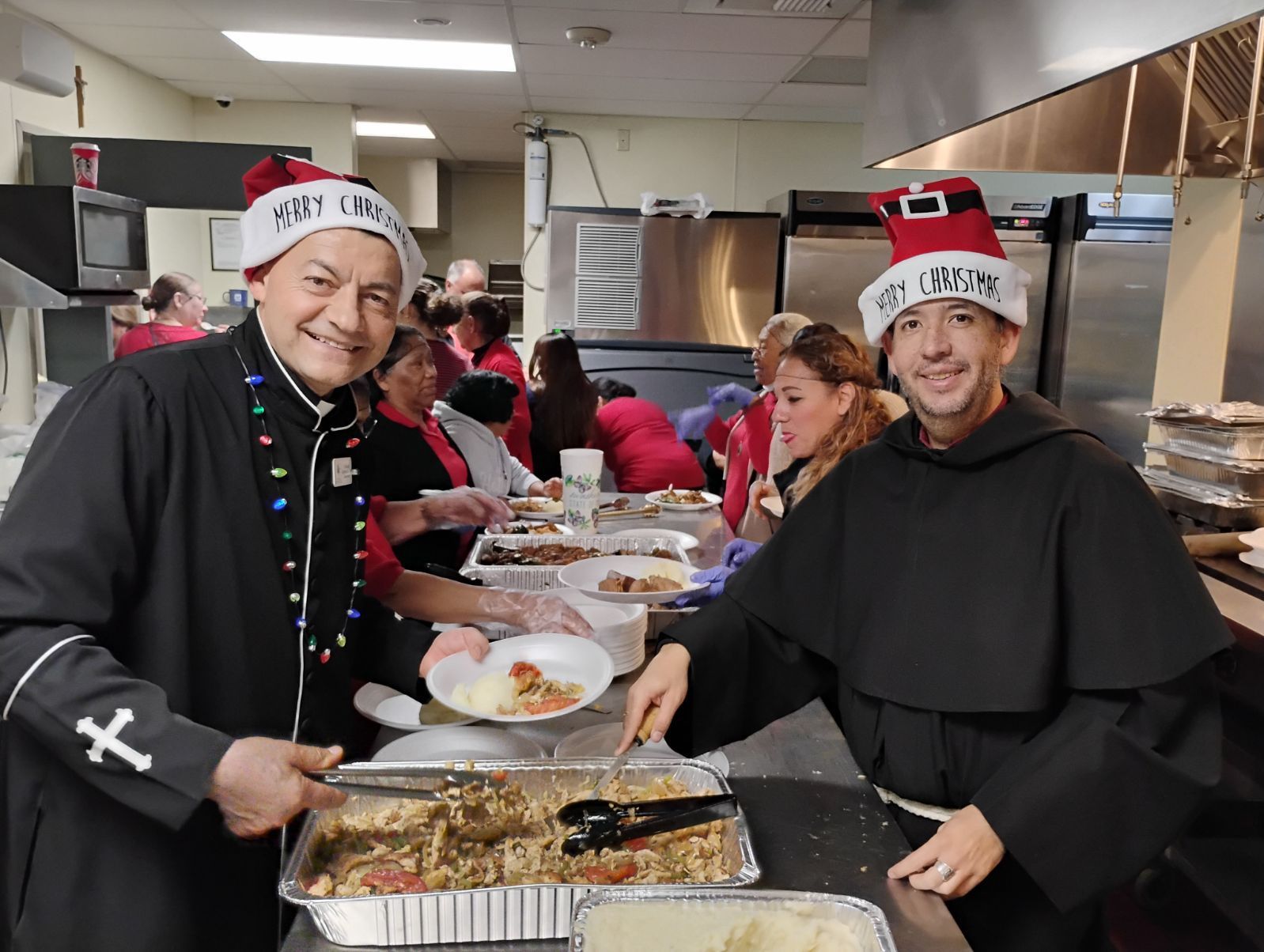 Two people in holiday hats serving food at a buffet in a kitchen