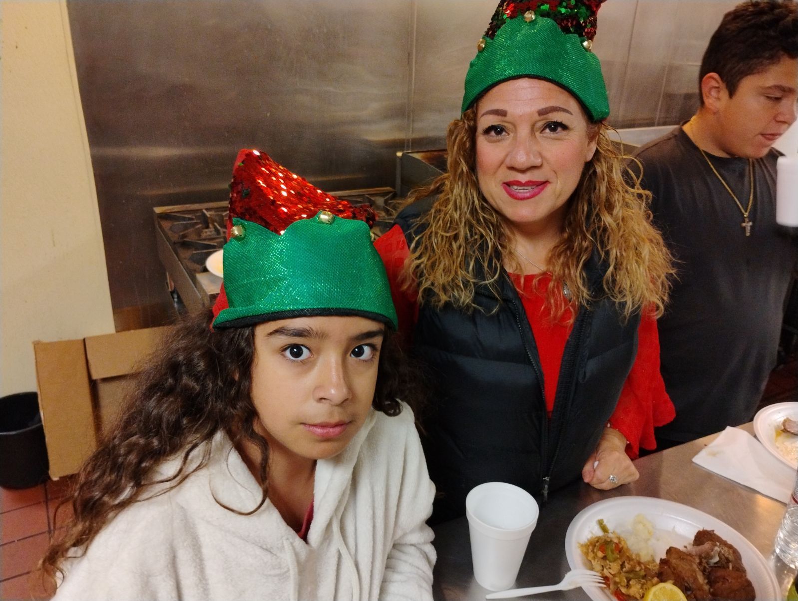 Two people in festive red-and-green hats sit at a table with plates of food and drinks.