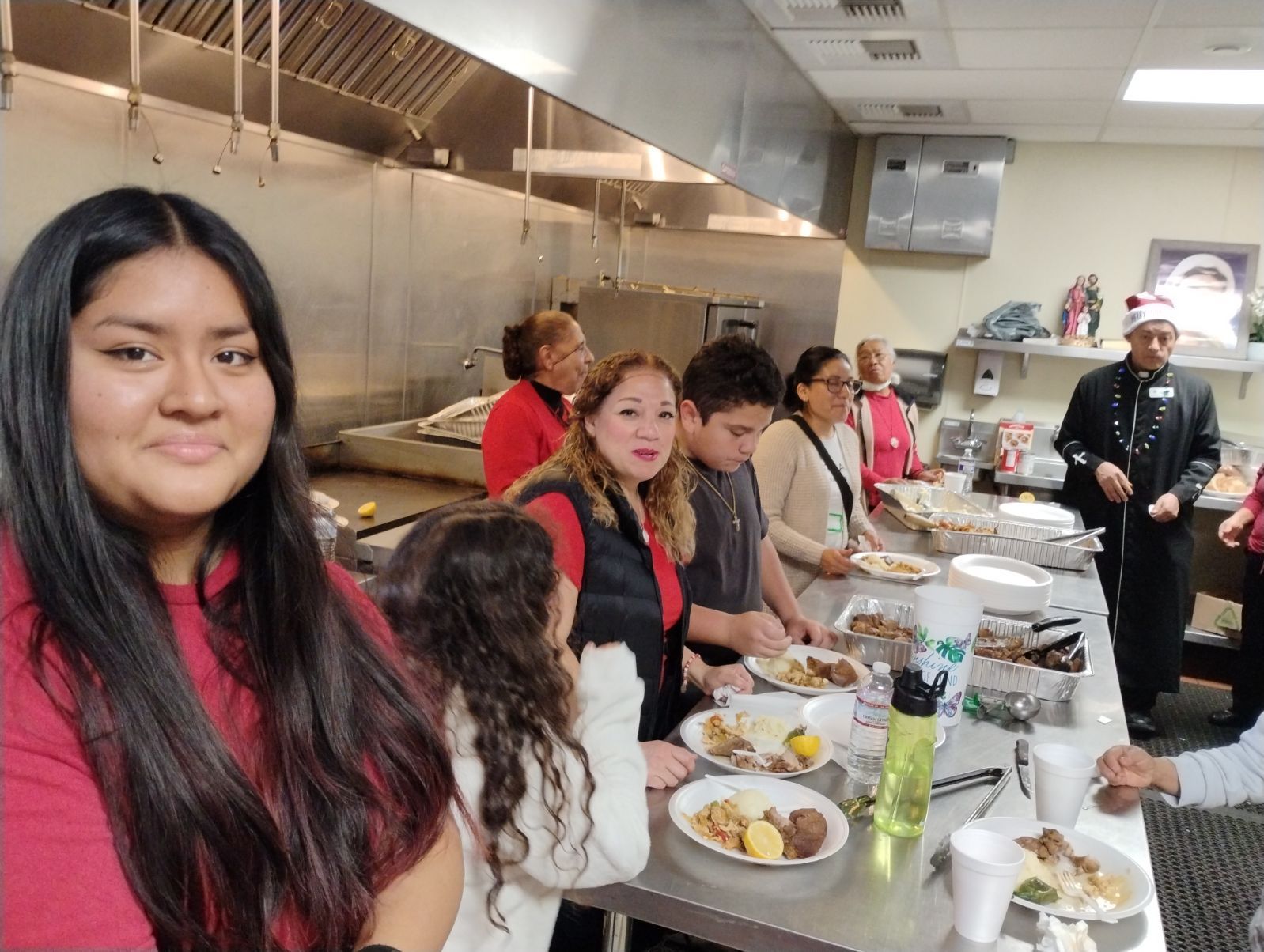 People seated at a long banquet table in a kitchen, sharing a meal and facing the camera.