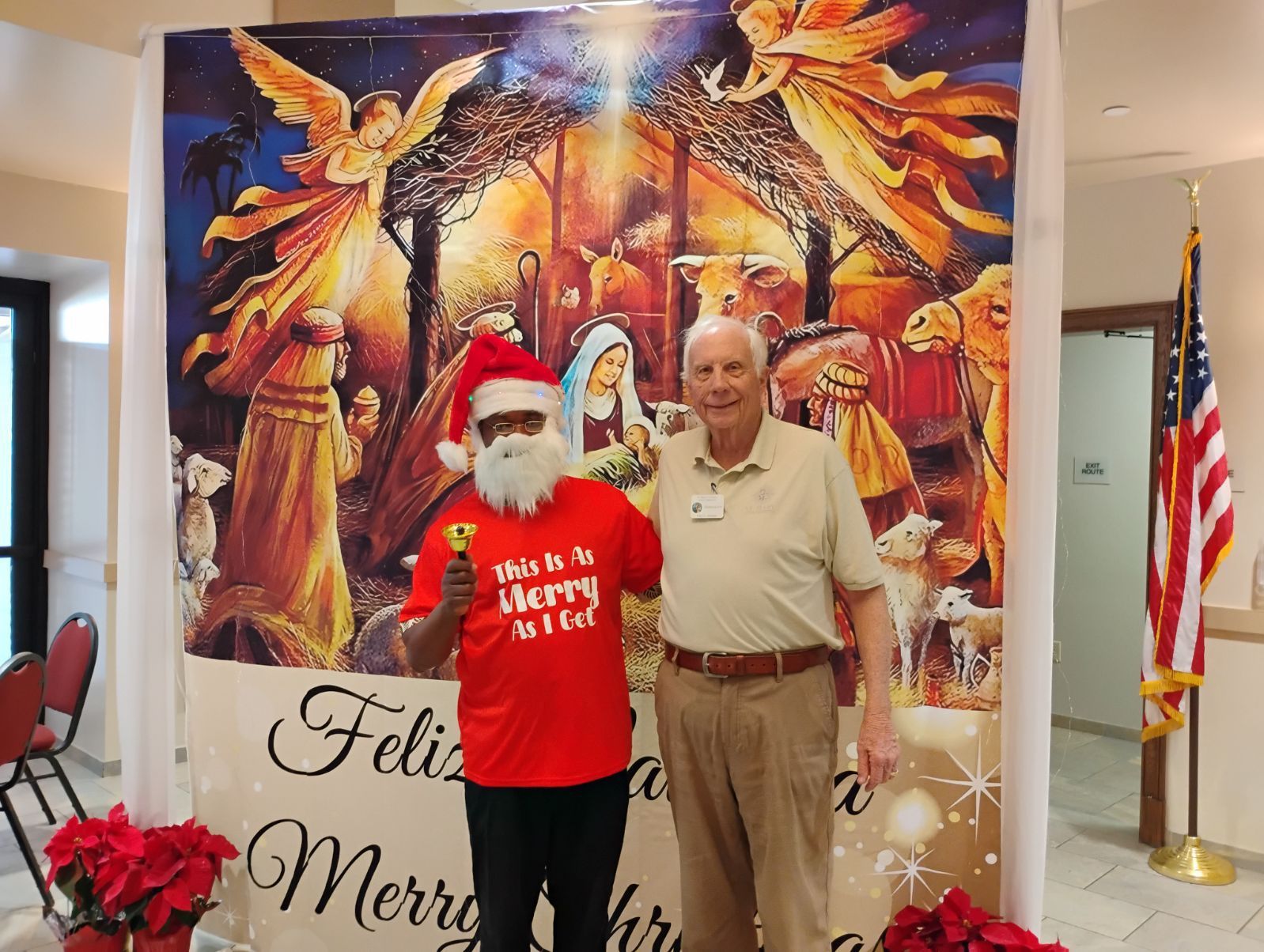 Two people posing at a Christmas display, one in a Santa hat and red shirt, with a festive mural and U.S. flag.