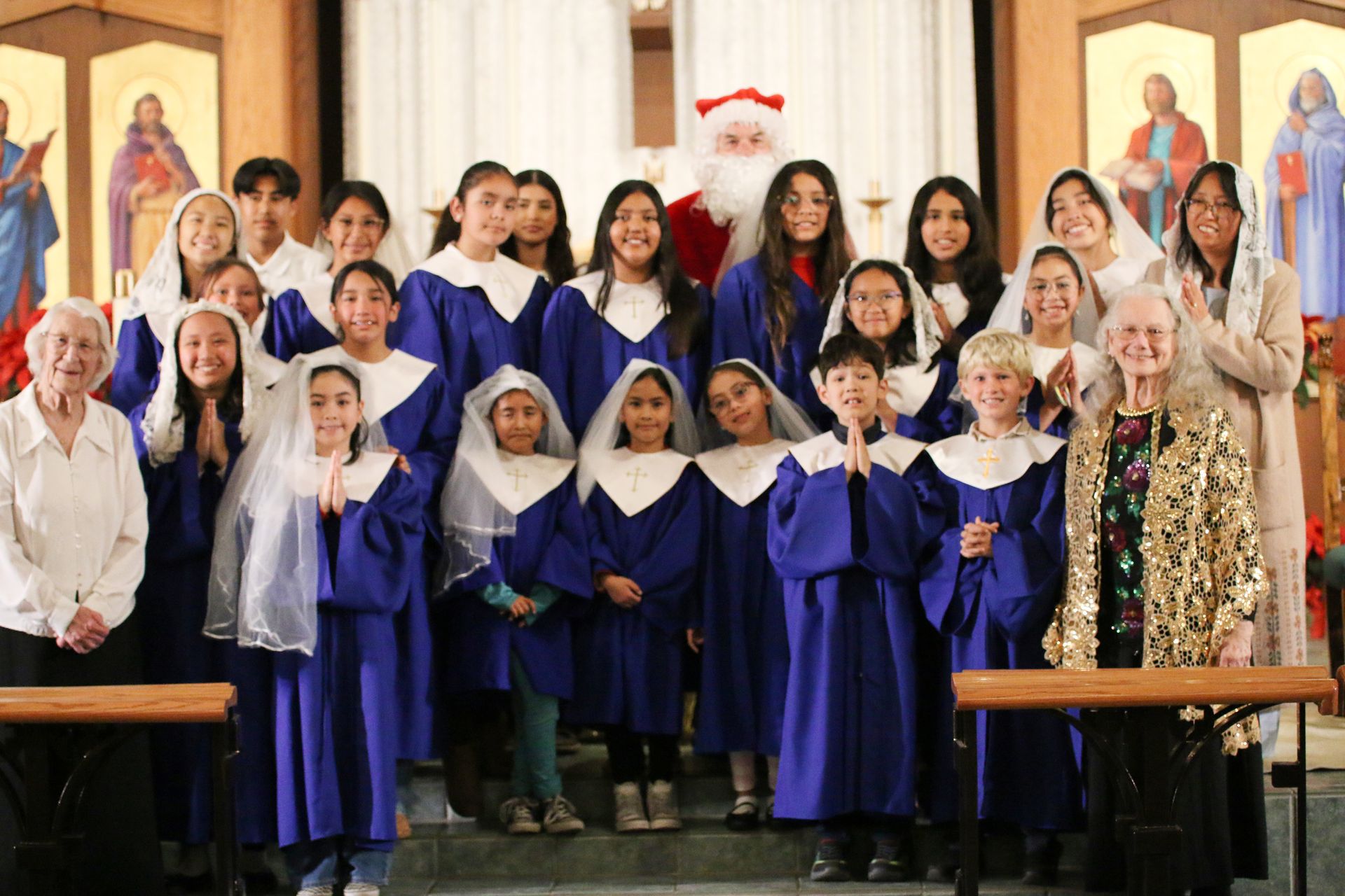 Choir group in blue robes standing in church, with a Santa figure and festive decorations behind them