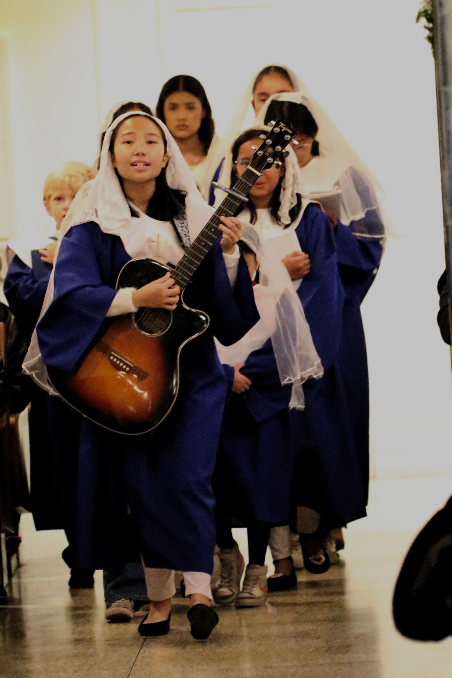 Women in blue robes walk in a procession, one playing a guitar-like instrument.