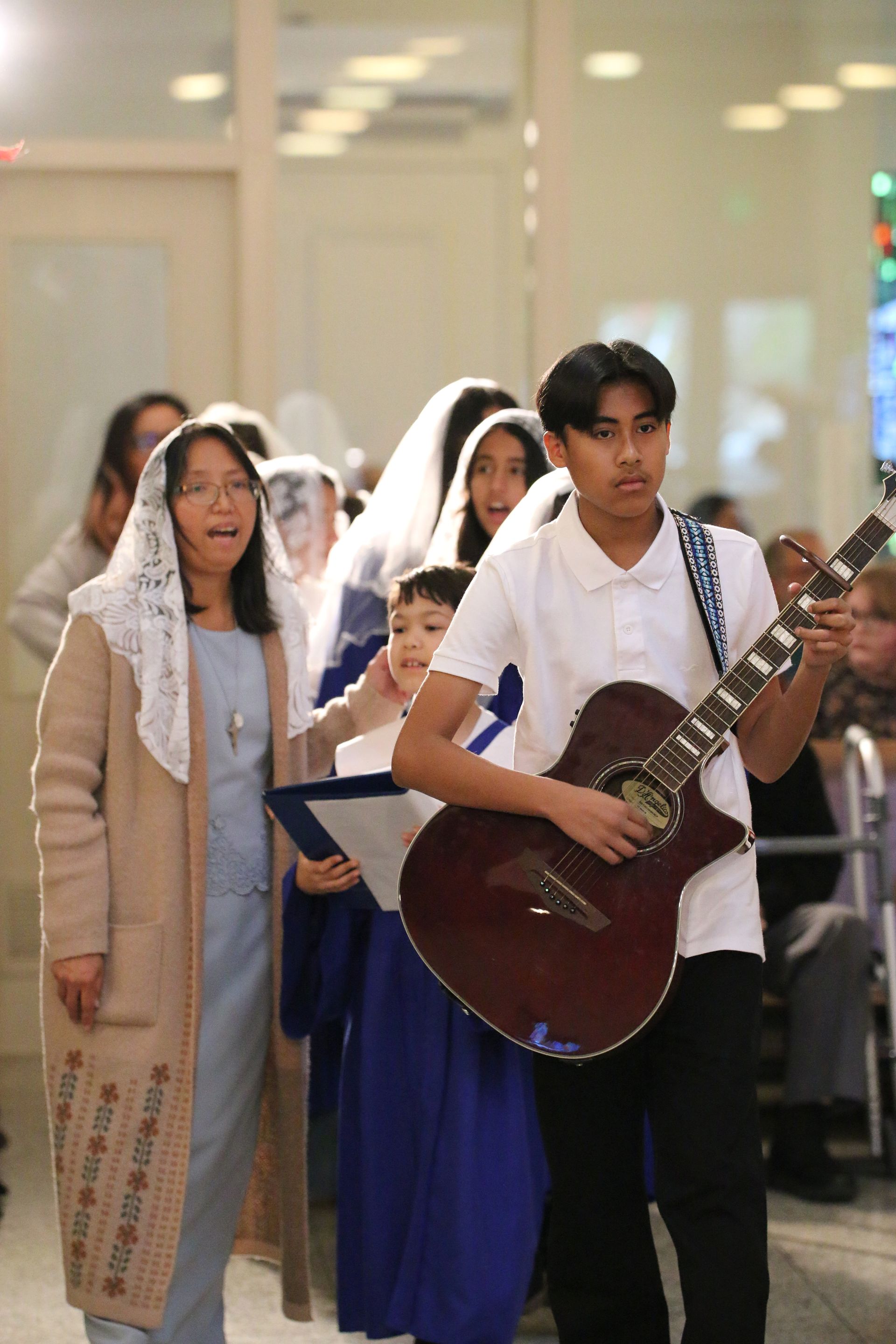 People in formal attire walking indoors, carrying guitars and sheet music in a procession.
