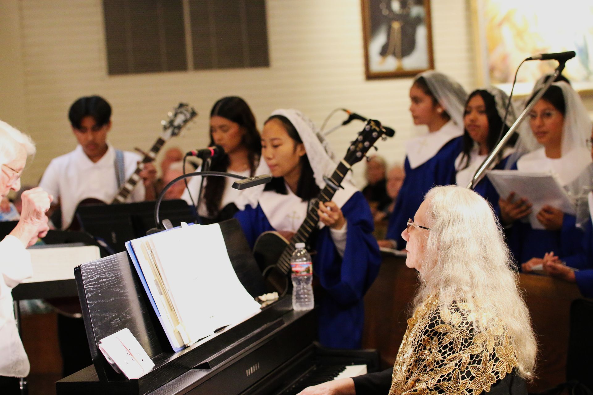 Choir rehearsing with guitars and sheet music around a piano in a church-like room