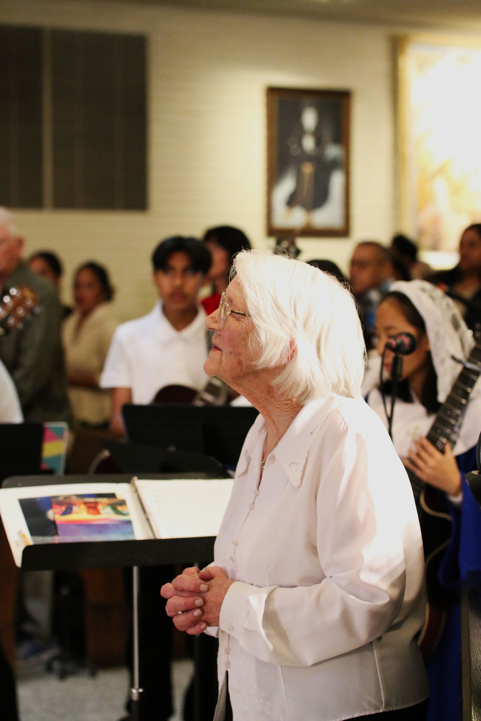 Woman in white headscarf standing in a meeting room, listening attentively with hands clasped.