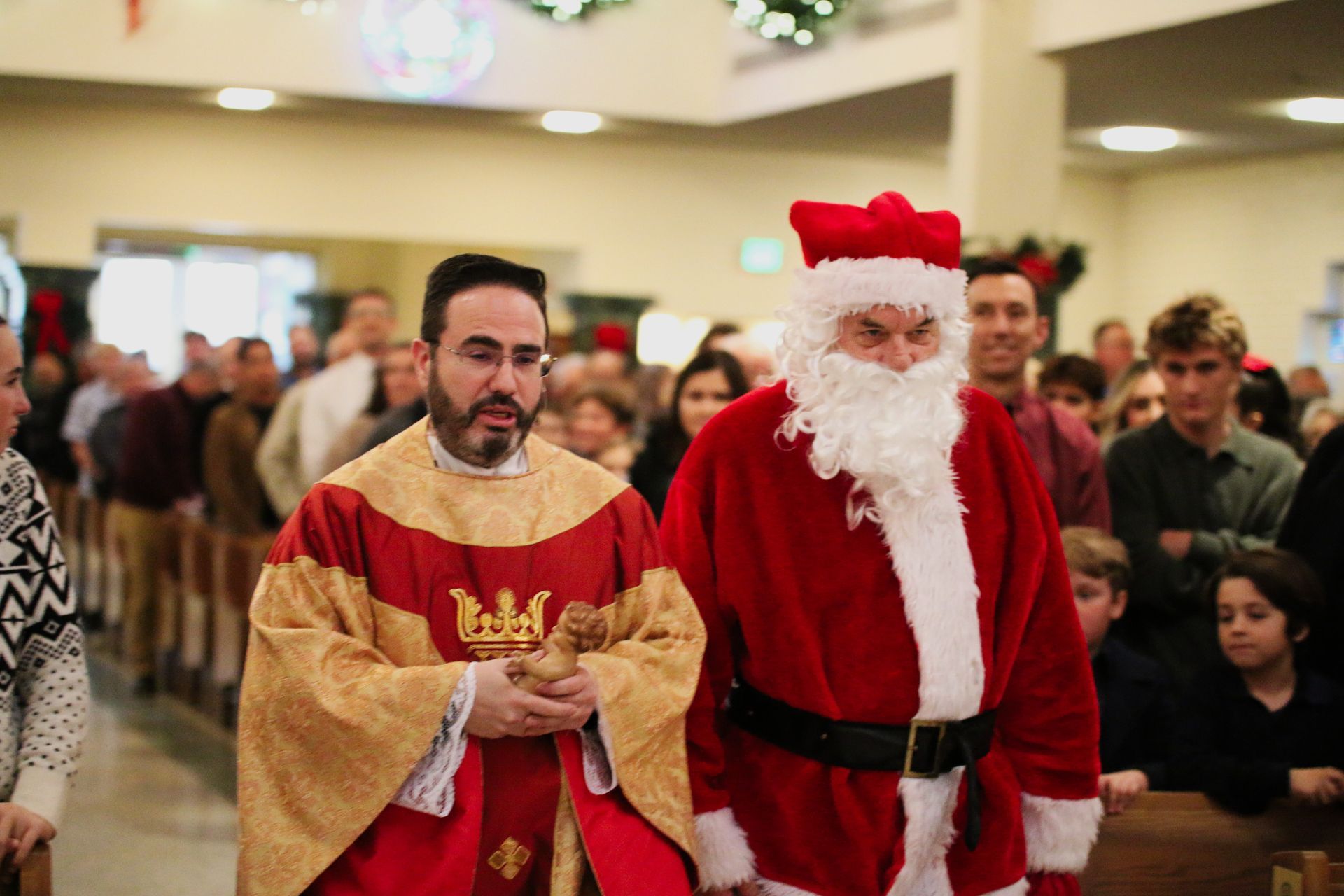 Santa in red suit walks beside a man in a gold costume through a crowded indoor event
