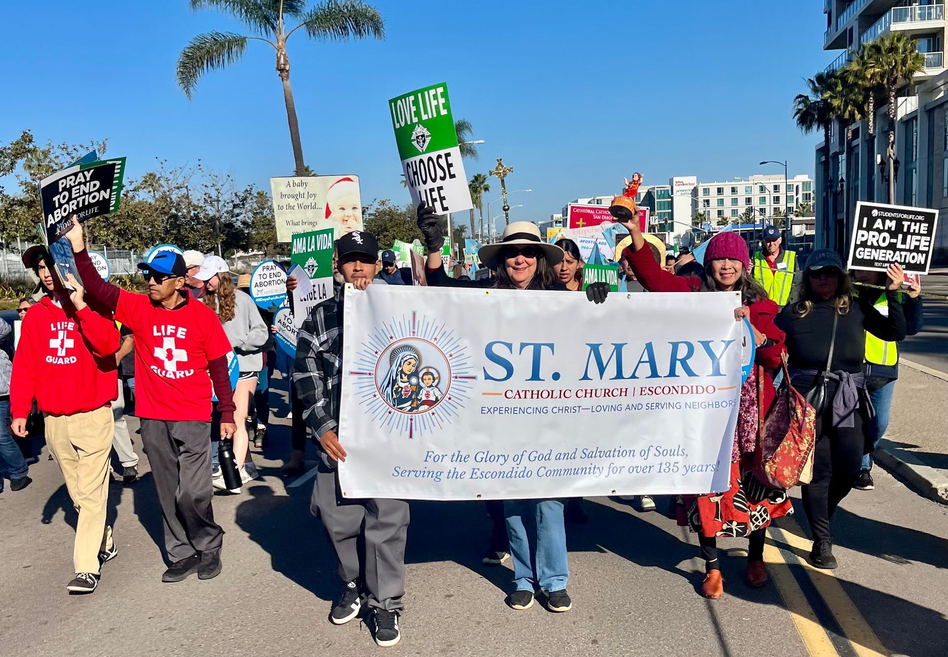 People marching in a sunny street protest holding a large St. Mary banner and signs, with palm trees nearby
