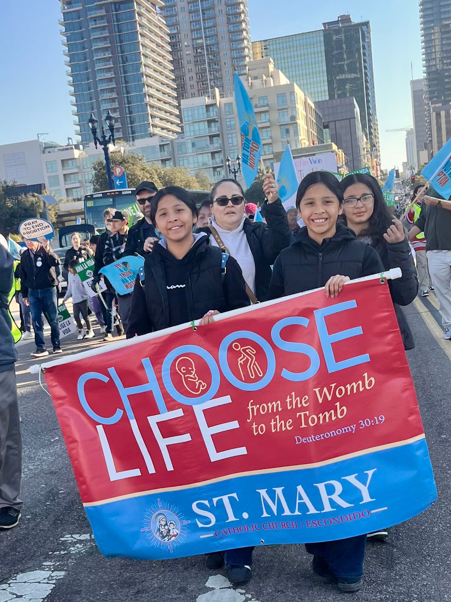 People holding a red “CHOOSE LIFE” banner at a city march, with buildings in the background.