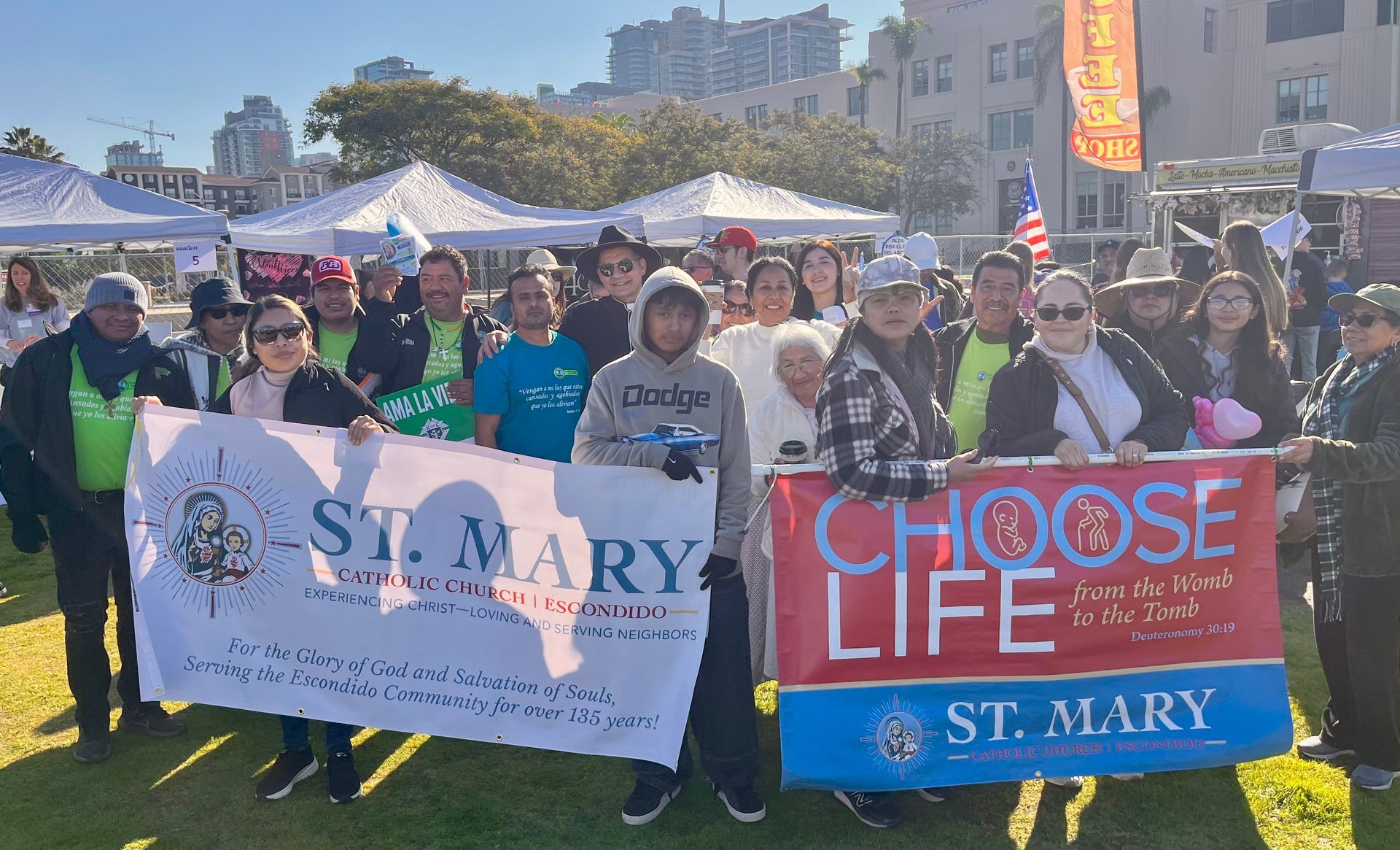 People holding St. Mary banners at an outdoor event with tents and a bright sky.