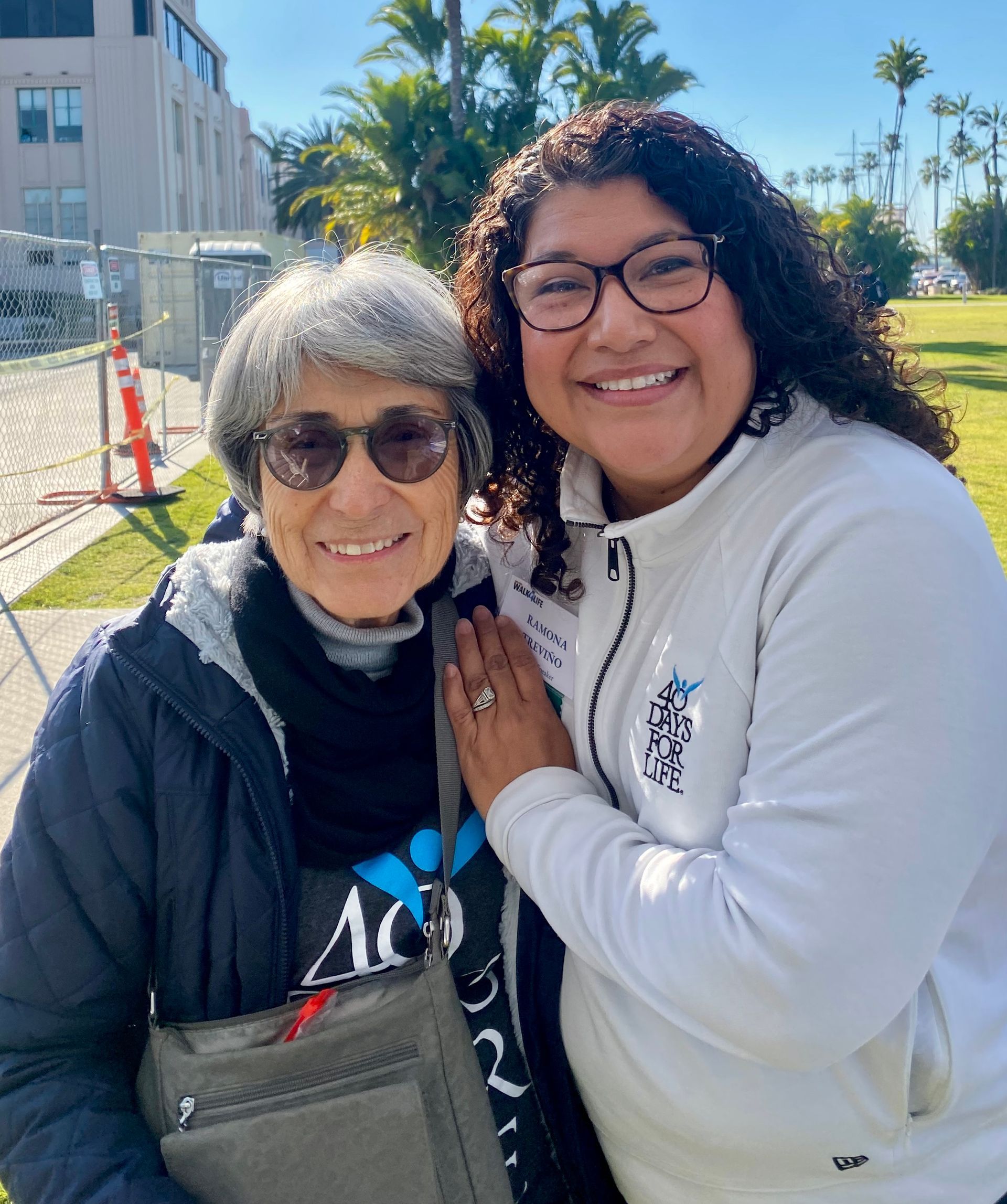Two smiling people posing outdoors in sunlight, one in a black jacket and one in a white hoodie.