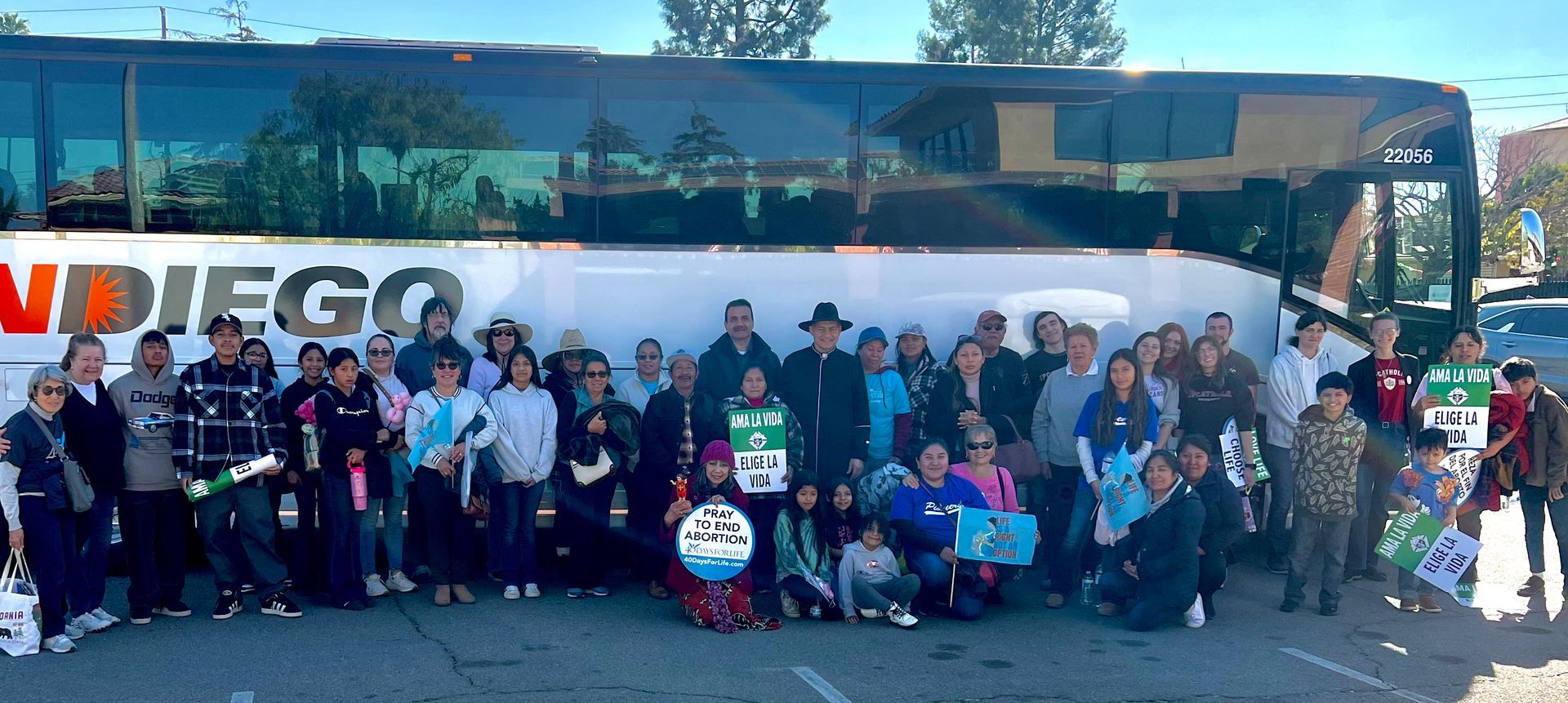 Large group posing in front of a blue coach bus with “SAN DIEGO” lettering, some holding signs and bags.