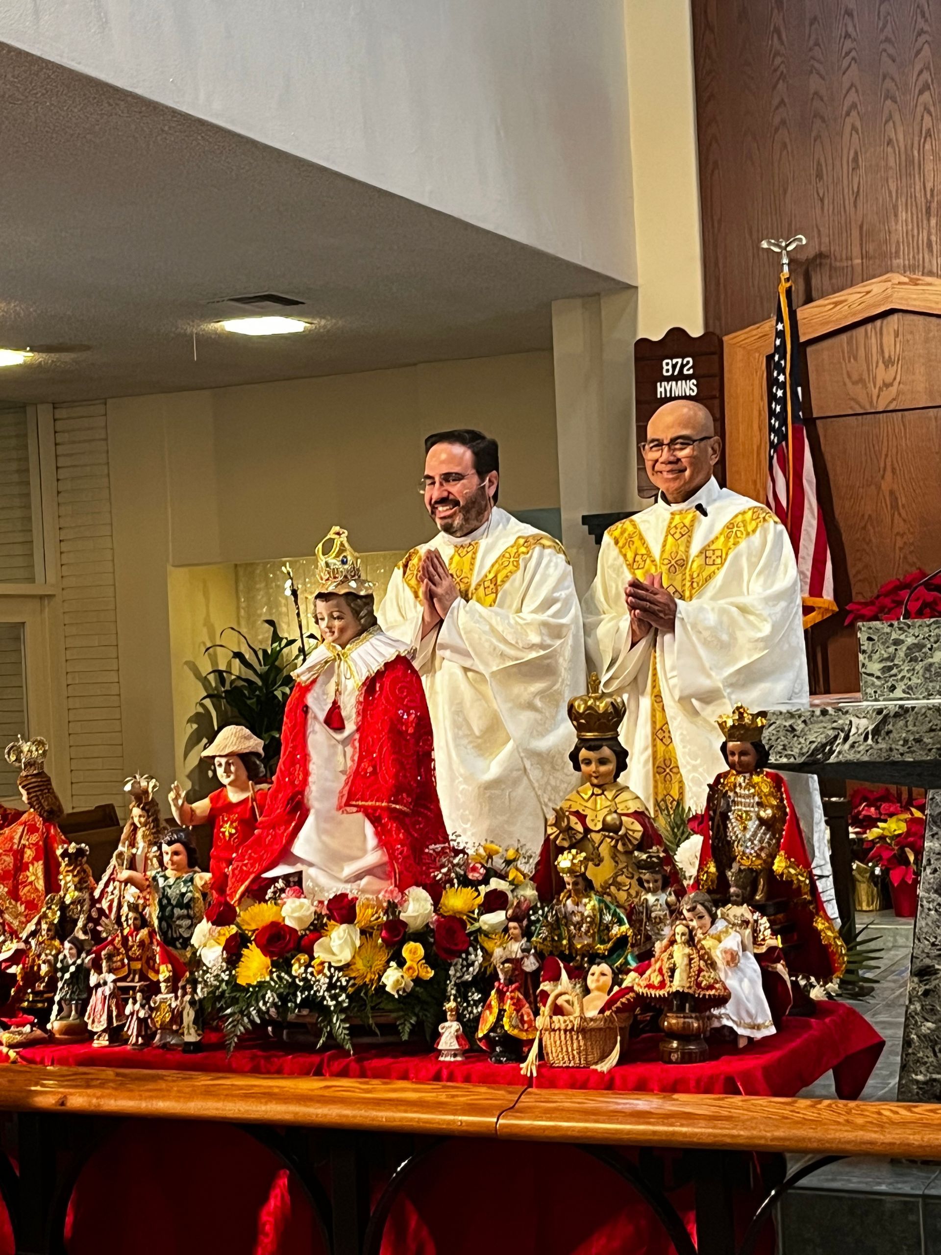Church altar with two priests in white and gold vestments beside a red-robed statue and flowers