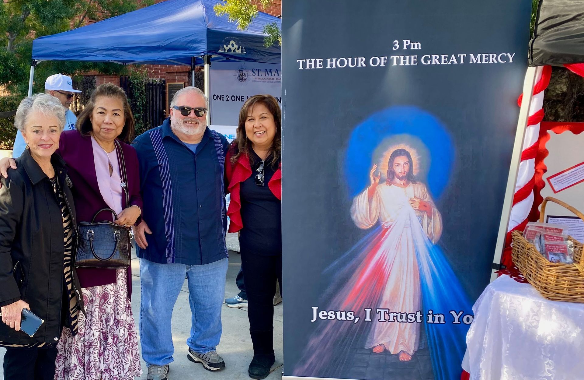 Group of people at an outdoor booth beside a banner reading “3 PM The Hour of the Great Mercy.”
