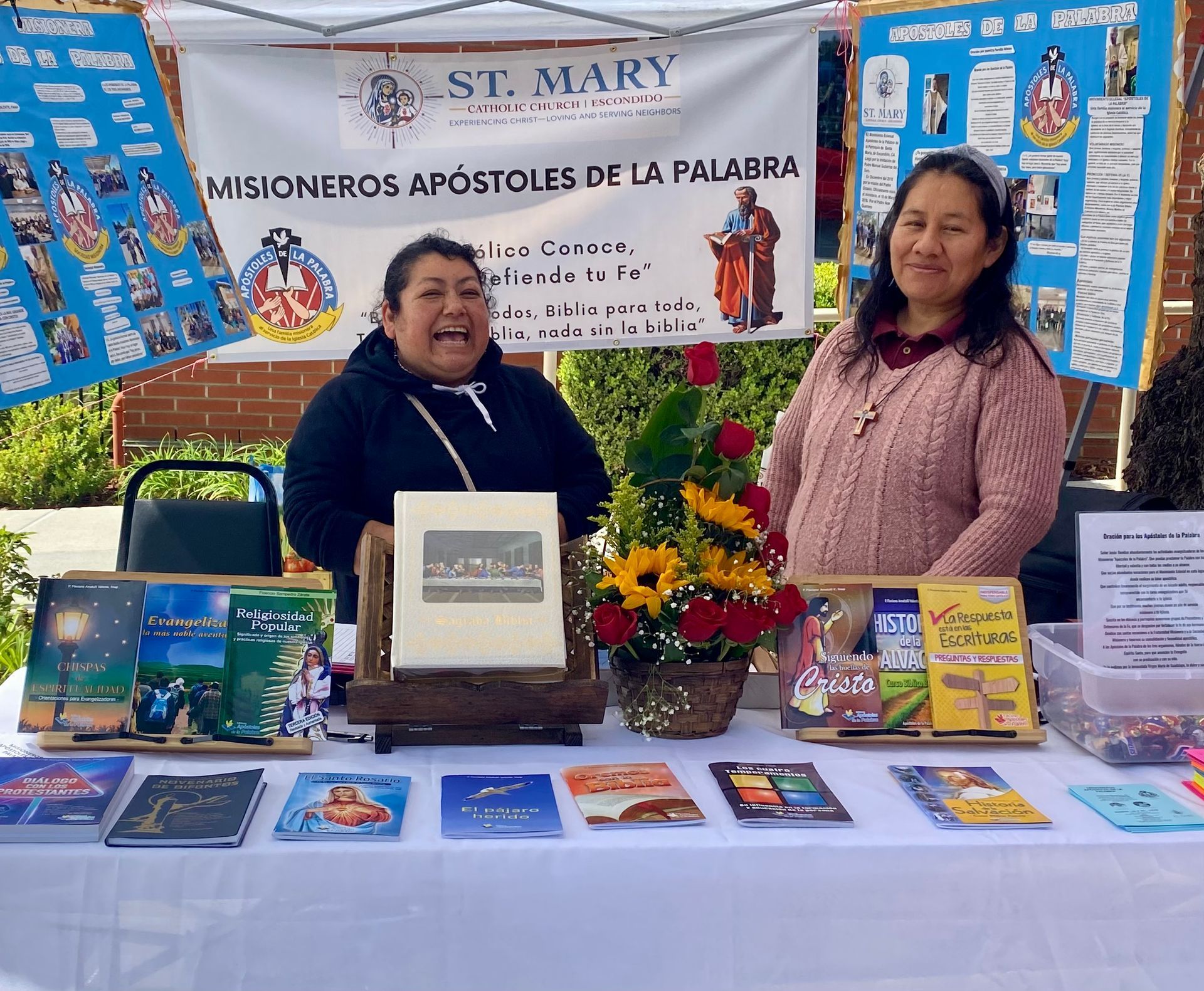 Two women at a St. Mary mission book table with religious books, banners, and display materials.
