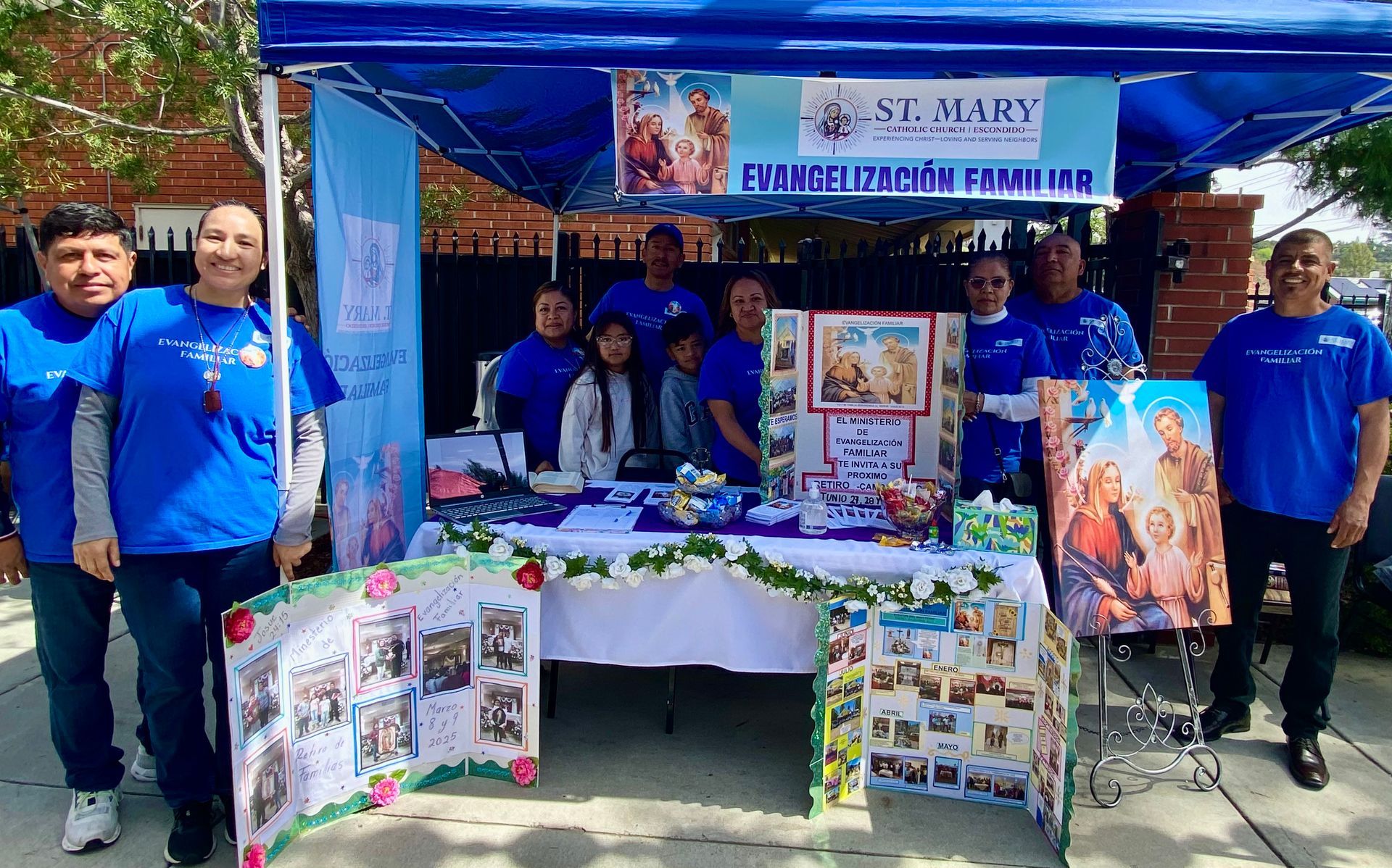 Group at a church booth with blue shirts, posters, and a banner reading “St. Mary” under a tent