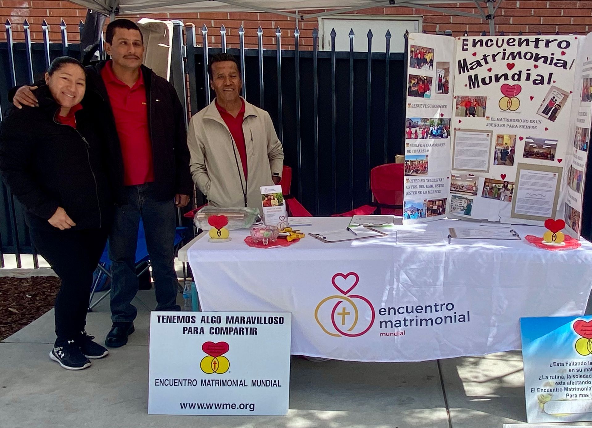 Three people stand beside a table with “Encuentro Matrimonial Mundial” display posters and brochures.