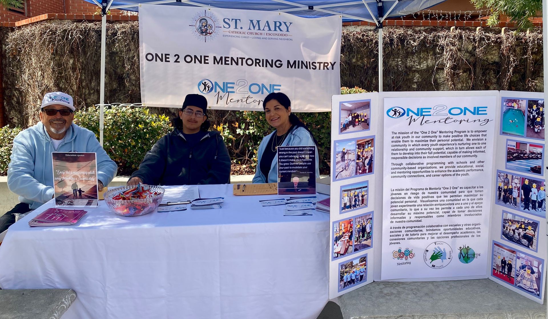 Three people at a white table display pamphlets and candy beside a “One One” informational booth.