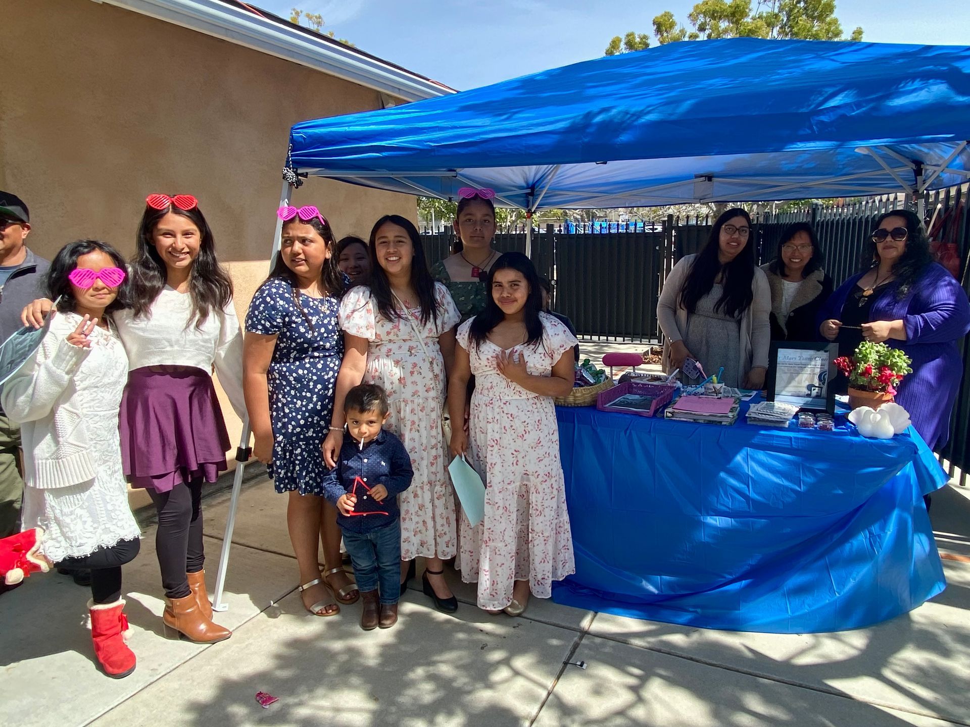 Group posing beside a blue canopy tent at an outdoor party table with gifts and decorations.