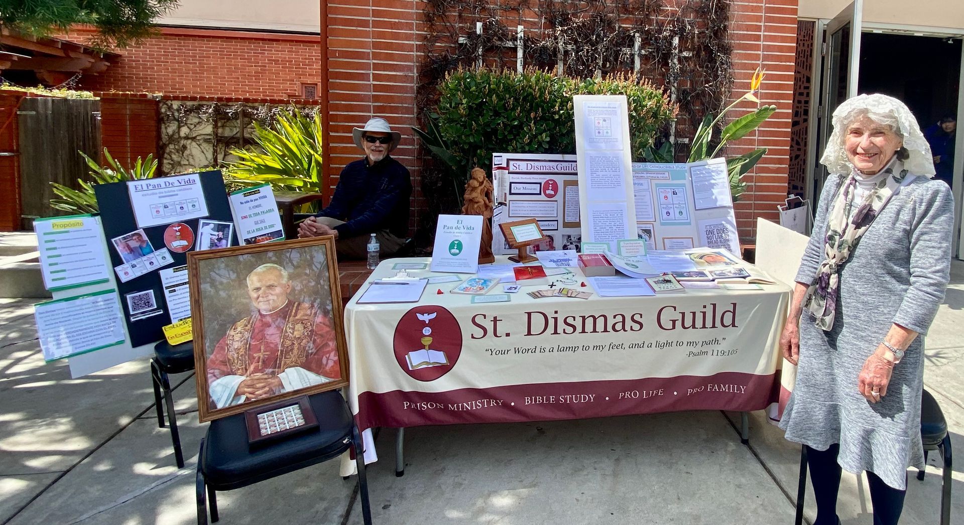 Outdoor St. Dismas Guild booth with posters, table display, and a woman standing beside it