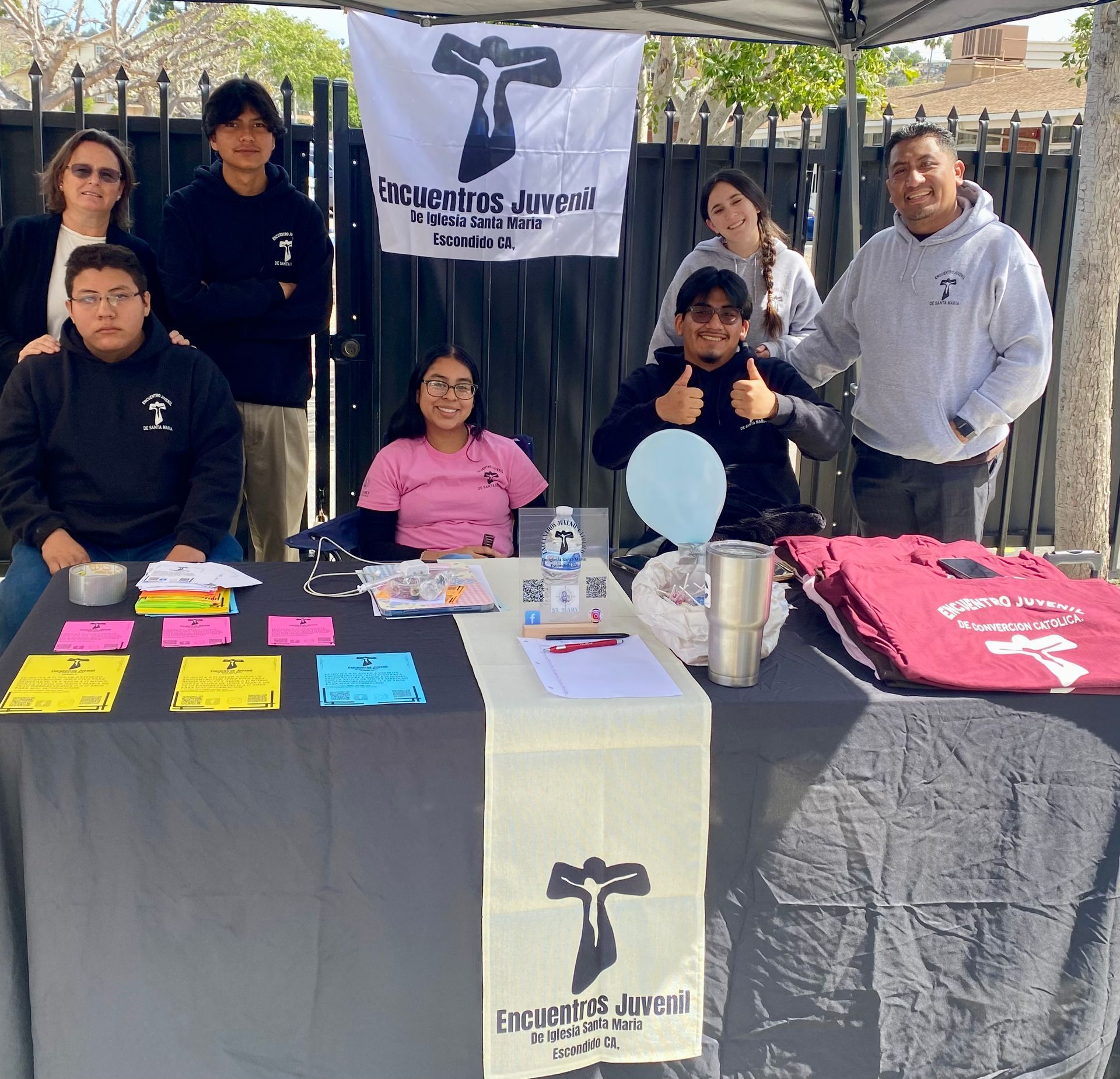 Volunteers at a campus outreach table with flyers, balloons, and a banner under a canopy