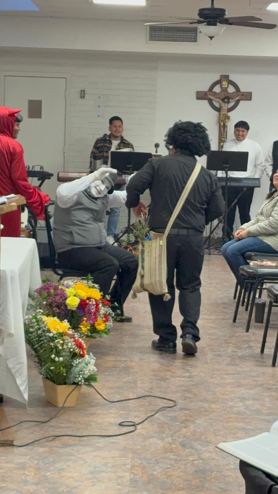 Musicians performing in a small church hall, with a floral arrangement in the foreground and audience seating nearby.