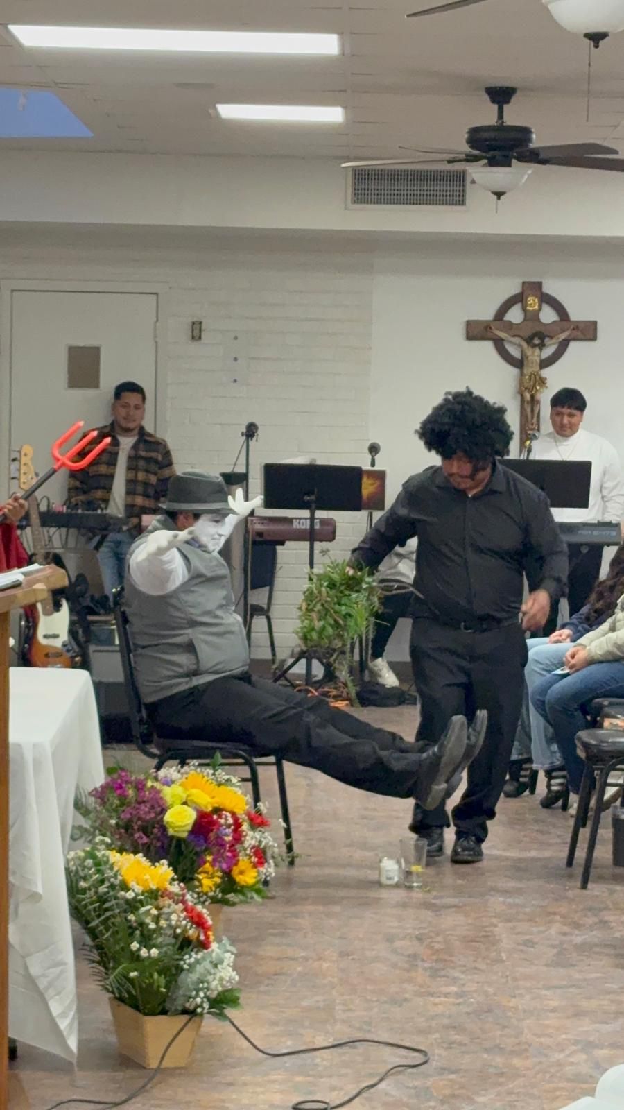 Person speaking in a small church hall with seated audience and floral arrangements near the front.