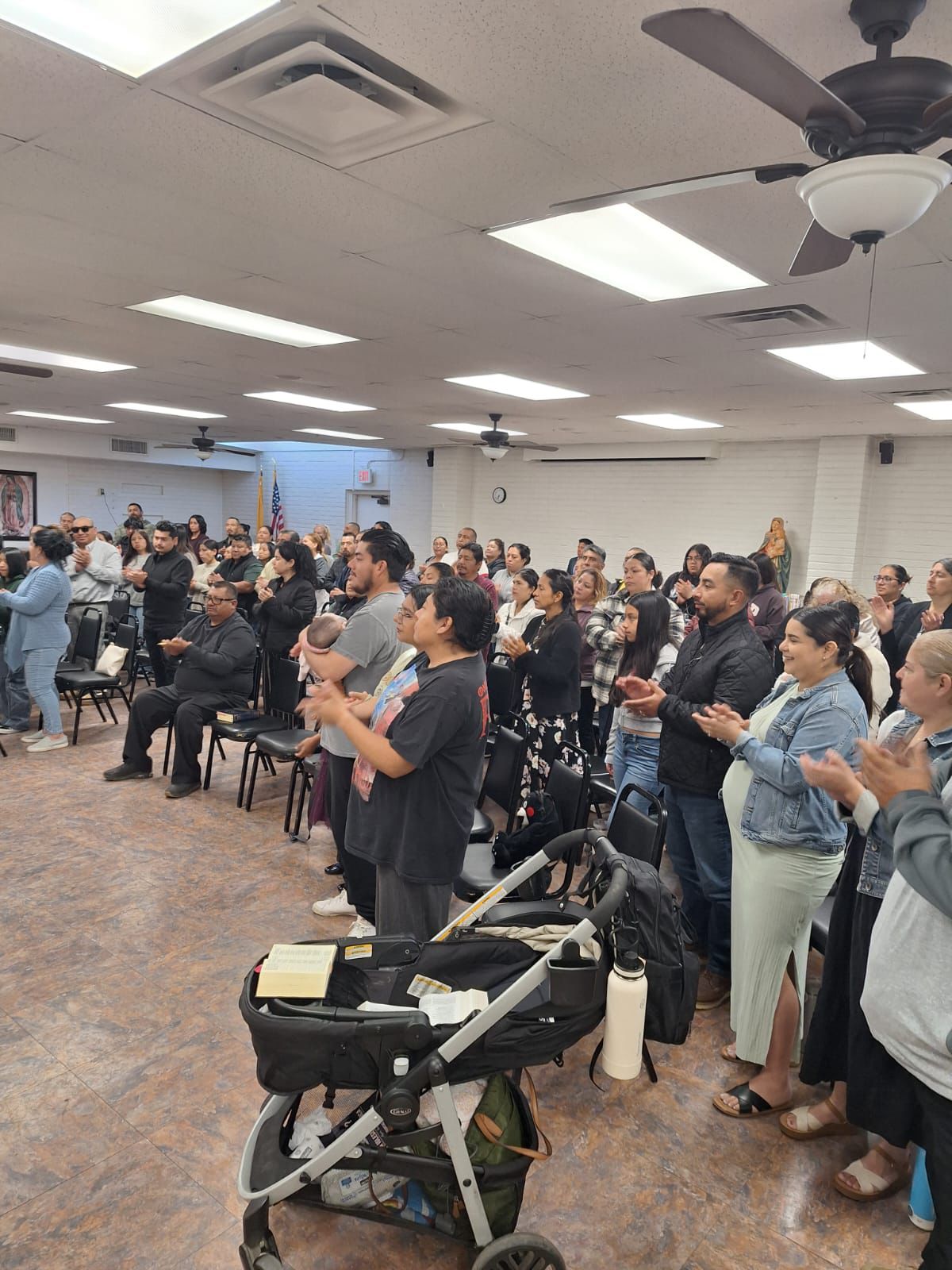 Audience standing and clapping in a conference room with rows of chairs and a stroller in the foreground