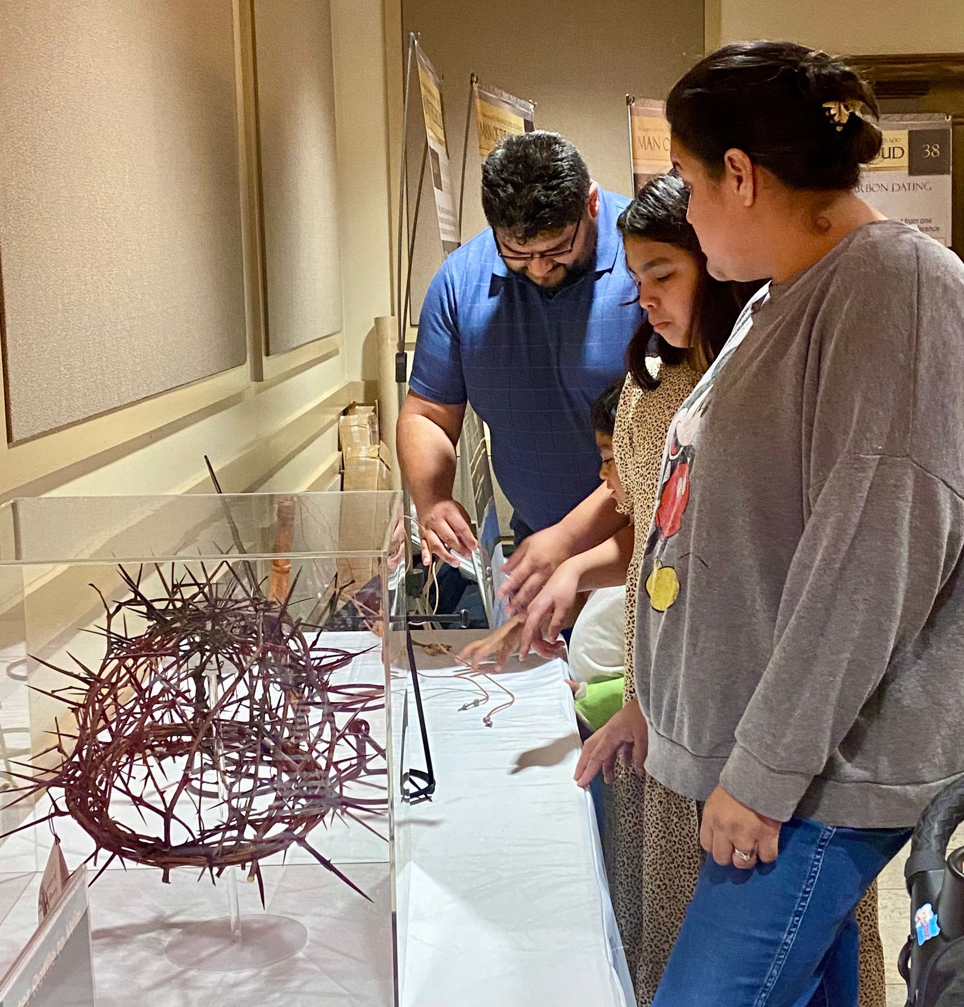 Three people examine a table display with a red wire sculpture in a gallery hallway.