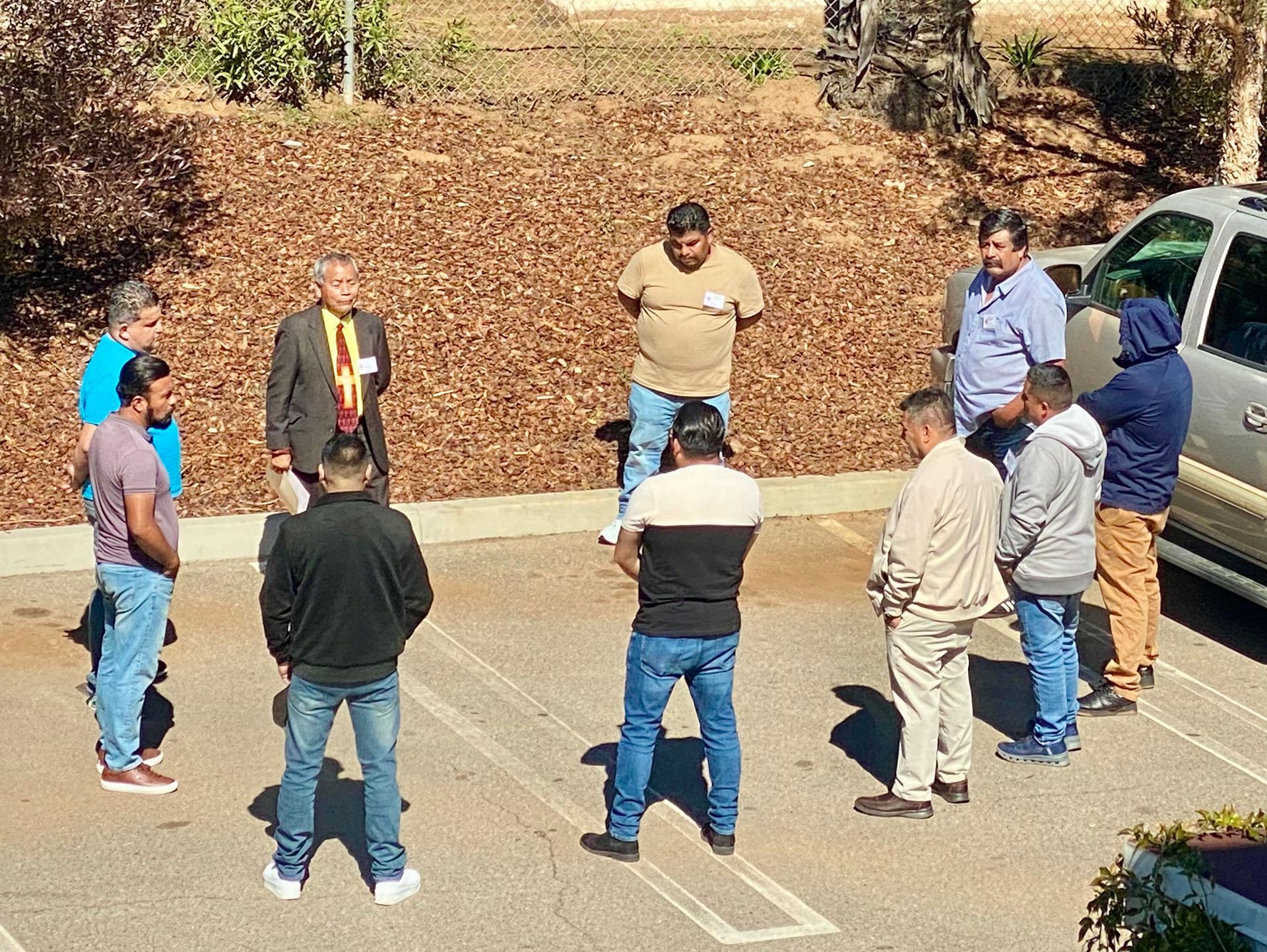 Small group standing in a circle outdoors on a road, talking near parked cars and dry leaves.