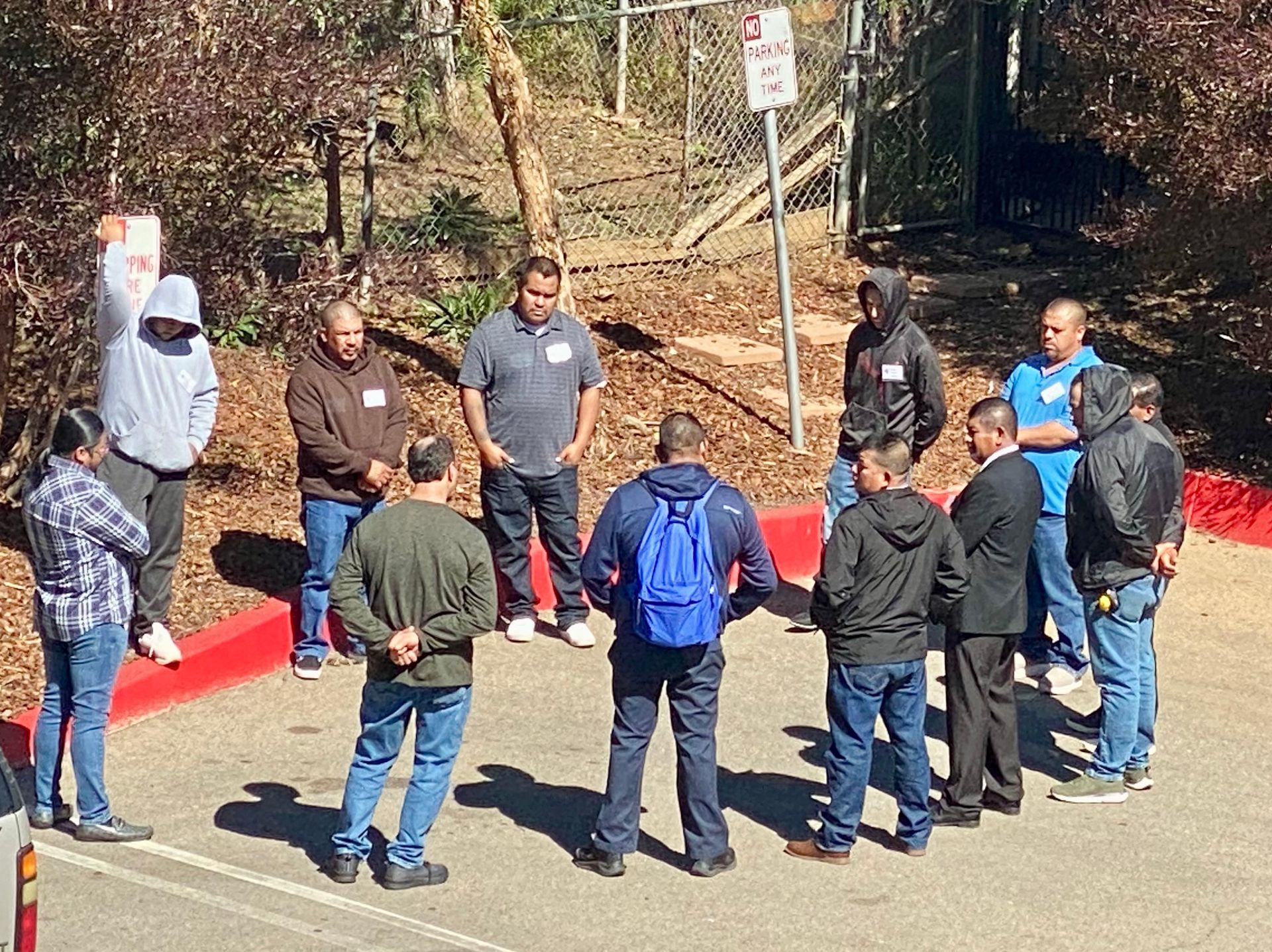 Group of people standing in a circle outdoors on a sunny roadside near wooded area