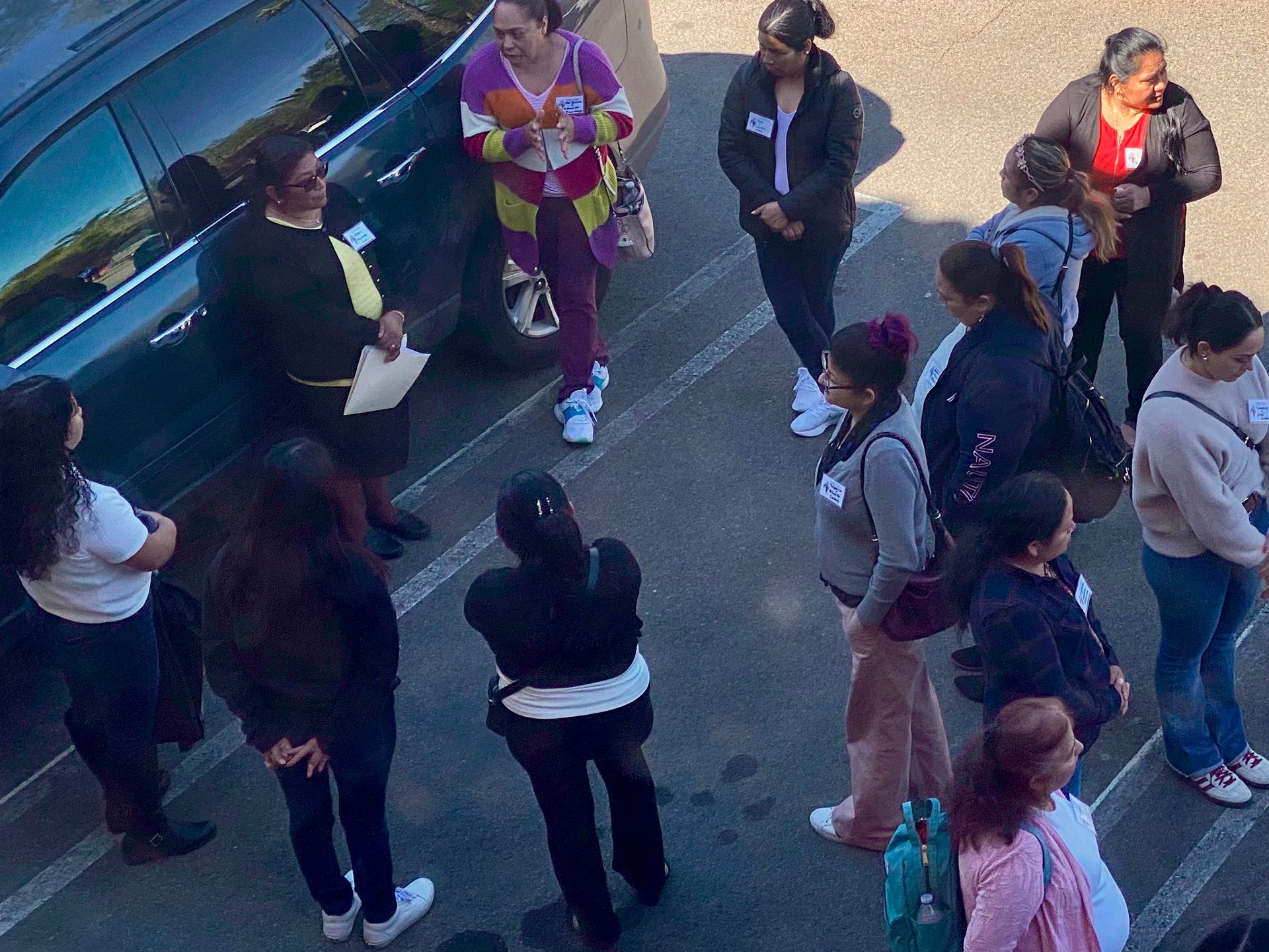 People standing in a parking lot near a blue vehicle, gathered in conversation.