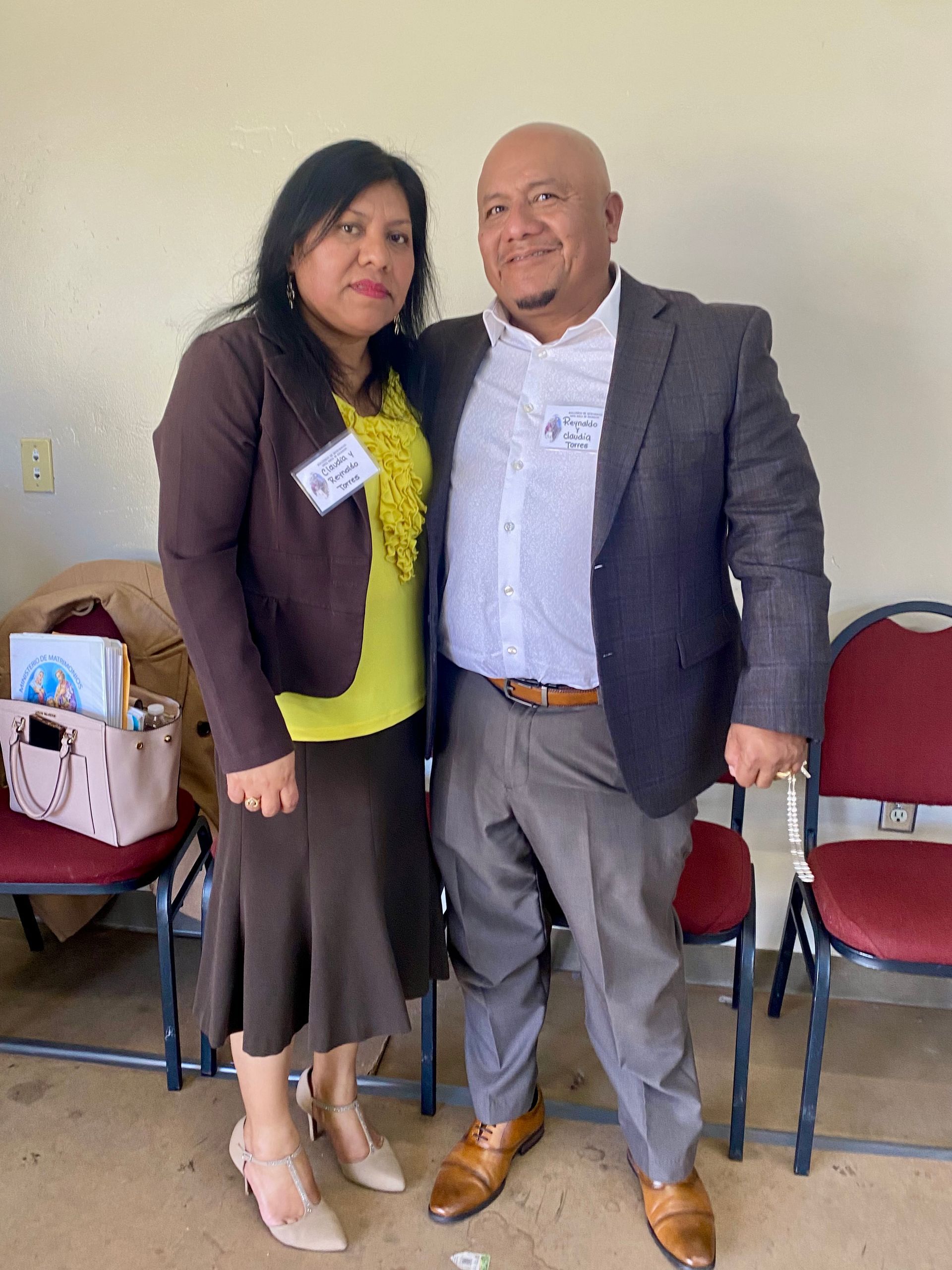 Two people standing together indoors, smiling, with chairs and bags in the background.