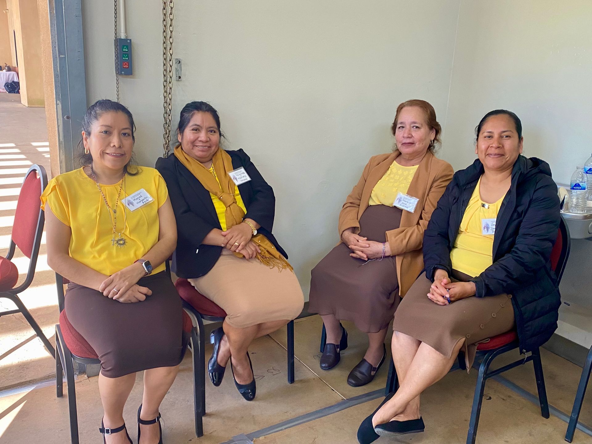 Four people seated indoors on chairs, wearing yellow shirts and dark outerwear, smiling at the camera.