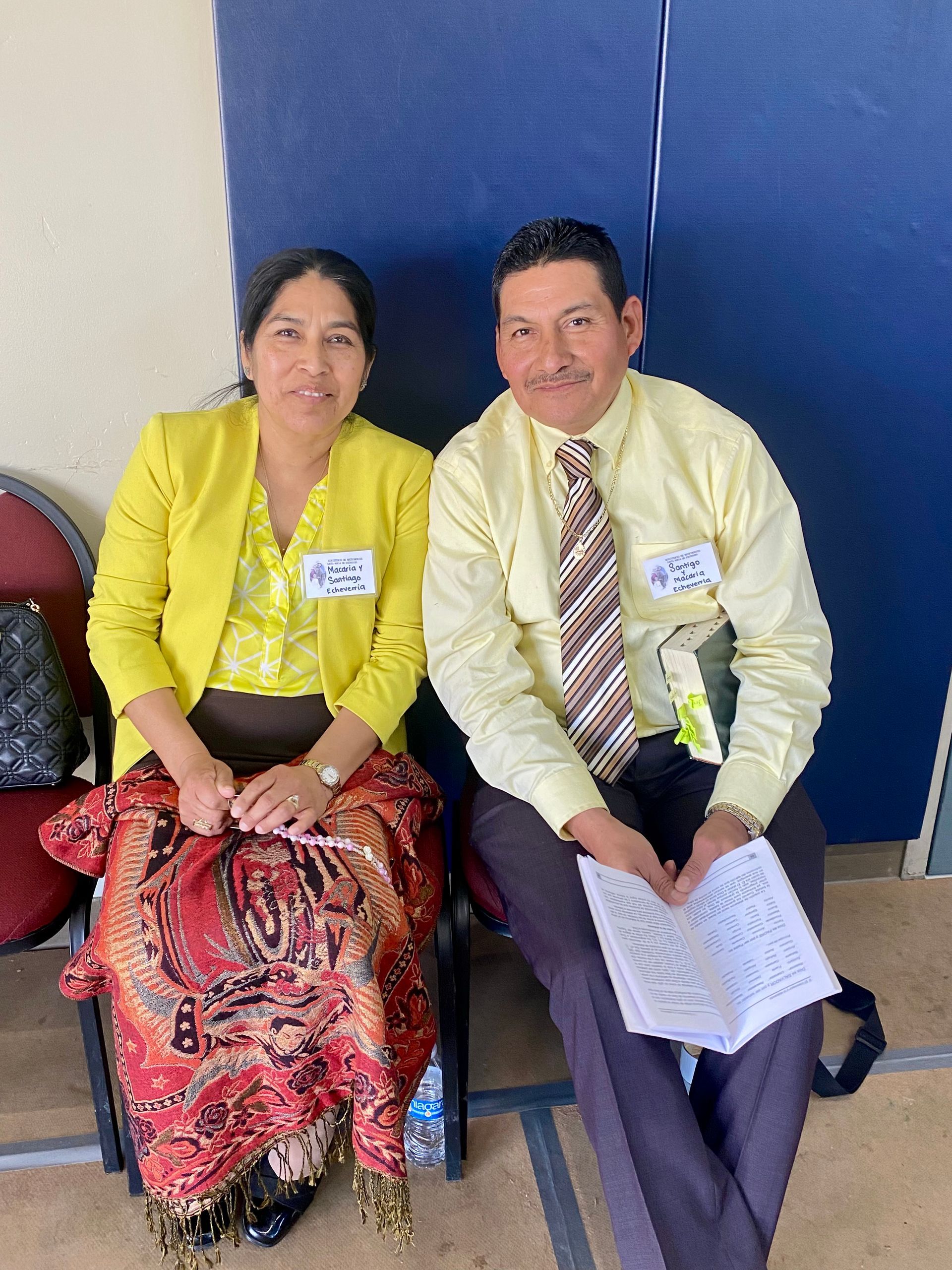 Two people seated side by side, wearing yellow tops and name tags, against a blue wall.