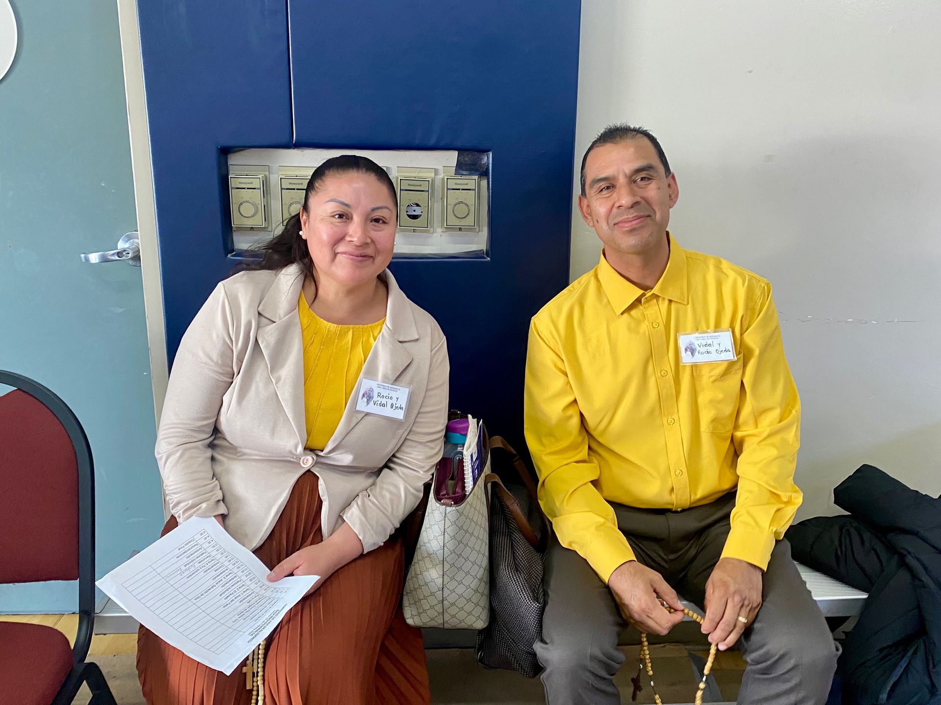 Two people seated indoors, one holding papers, wearing beige and yellow shirts.