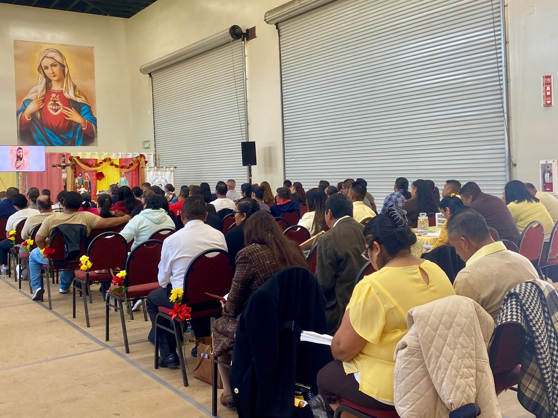 Audience seated in a hall facing a stage with a colorful portrait and speakers on the front wall