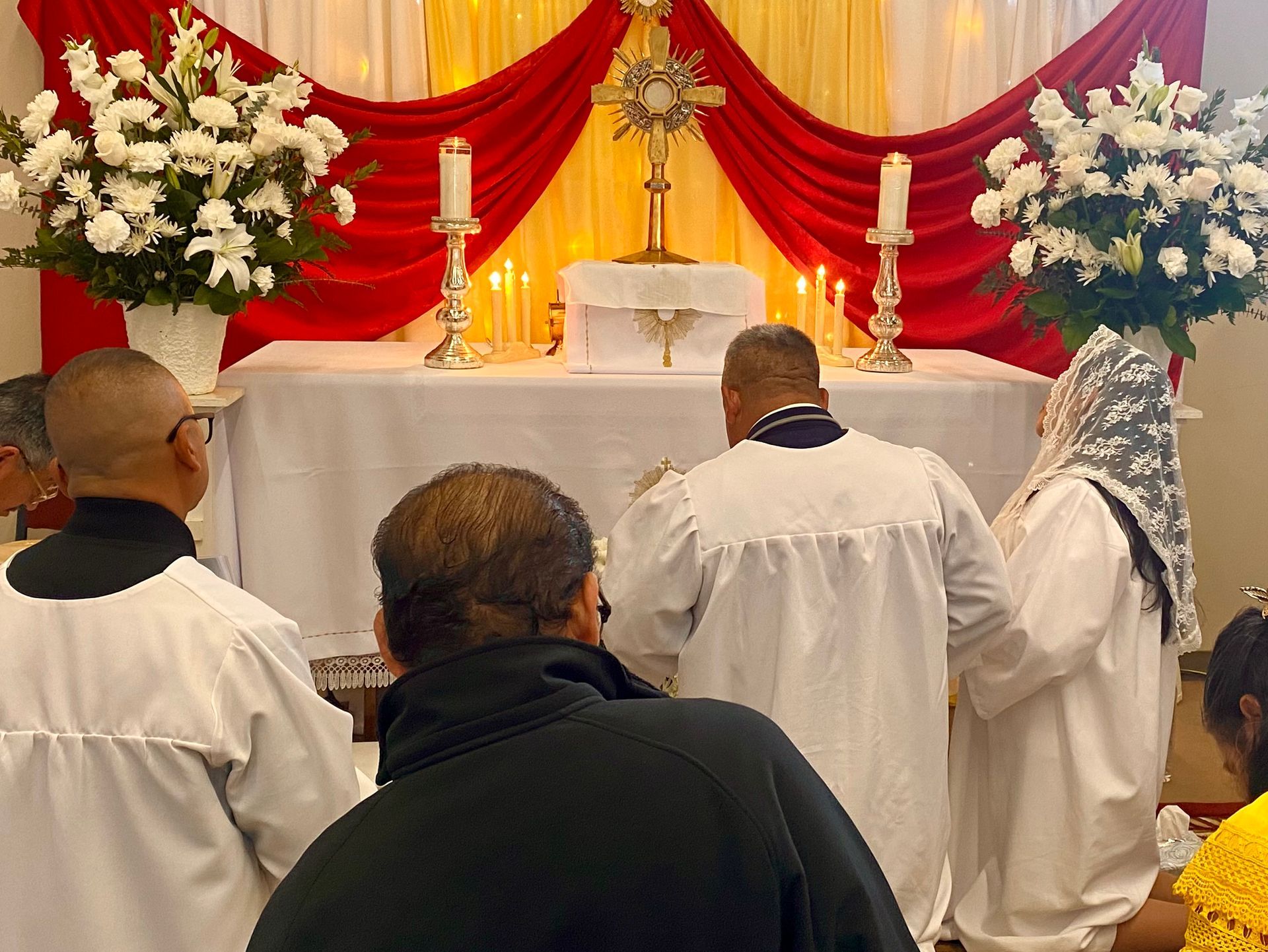 People in white robes kneel before a candlelit altar with red and cream drapes and floral arrangements.