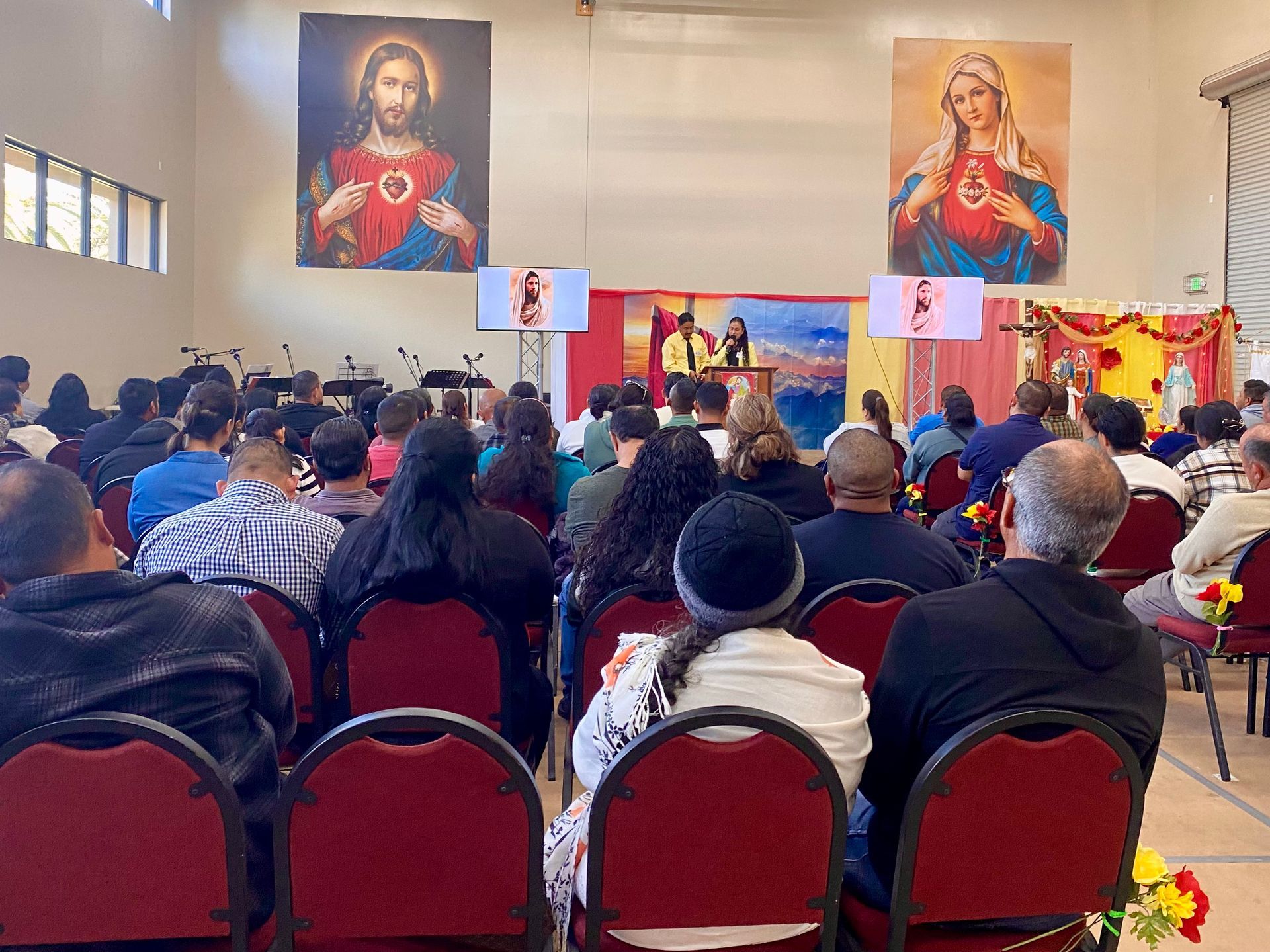 Congregation seated in a church hall facing a speaker and religious banners beneath large sacred portraits