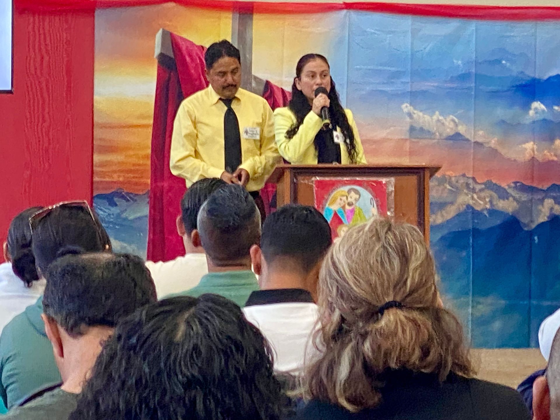Speaker at podium addressing audience, with another man beside him in front of a mountain backdrop.