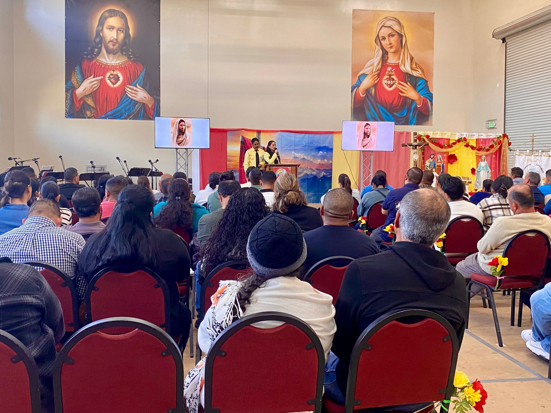 Congregation seated in a church hall facing a stage with religious banners and large sacred portraits.