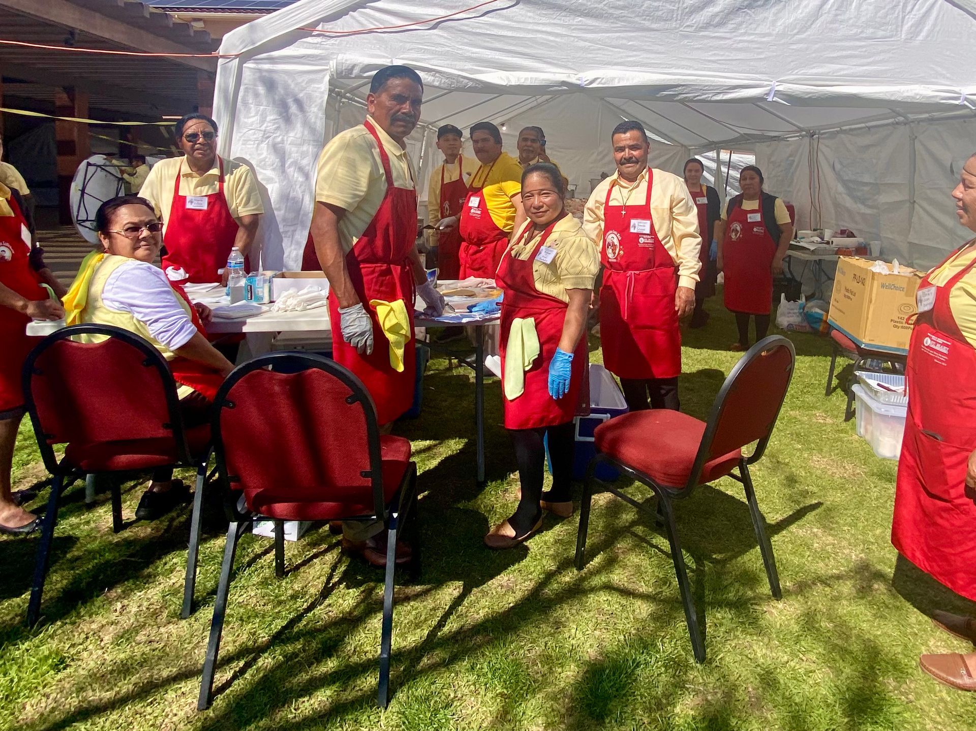 People in red aprons standing and sitting around chairs under a white tent on a grassy field