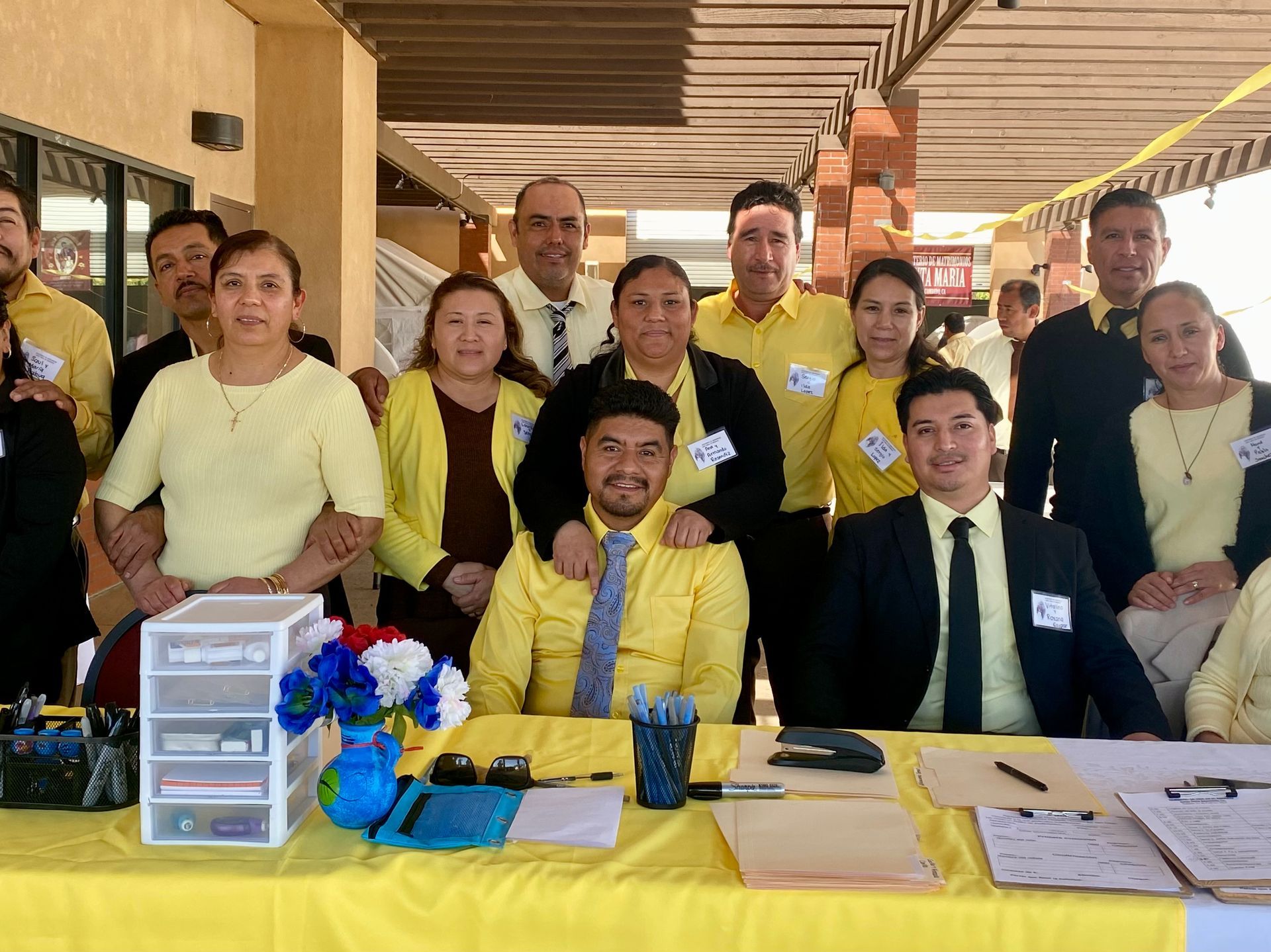 Group of people posing at a table with yellow and white flowers and documents in a covered outdoor setting