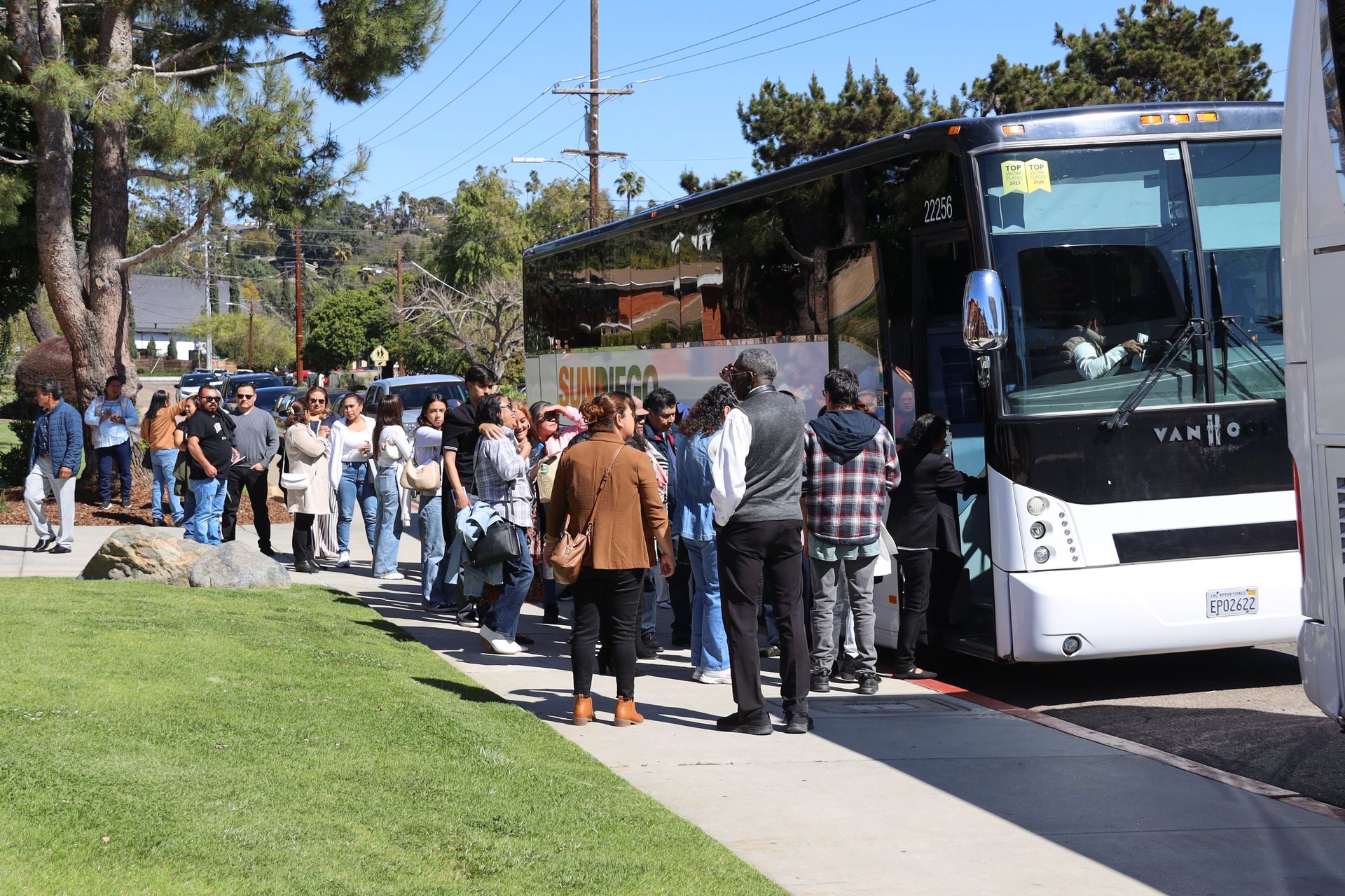 People waiting in line to board a white bus along a sunny sidewalk.