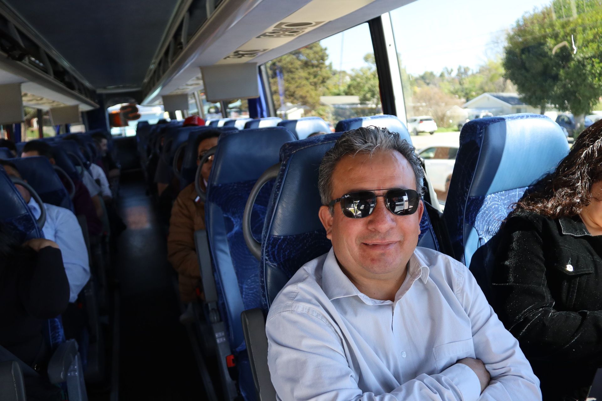 Man in sunglasses smiling on a bus, seated among other passengers in daylight.