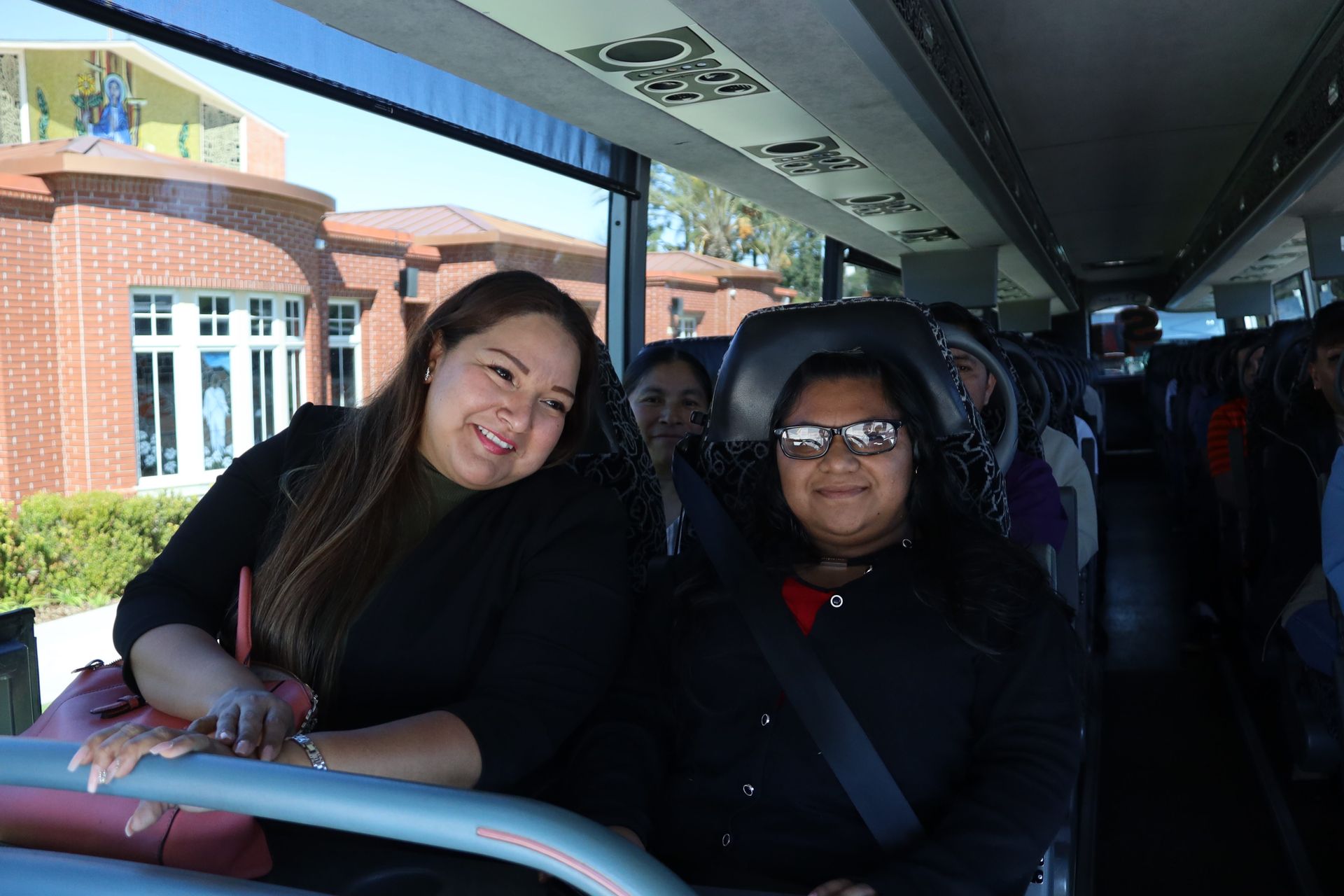 Two people smiling on a bus, one leaning on the seat rail with a school building outside the window.