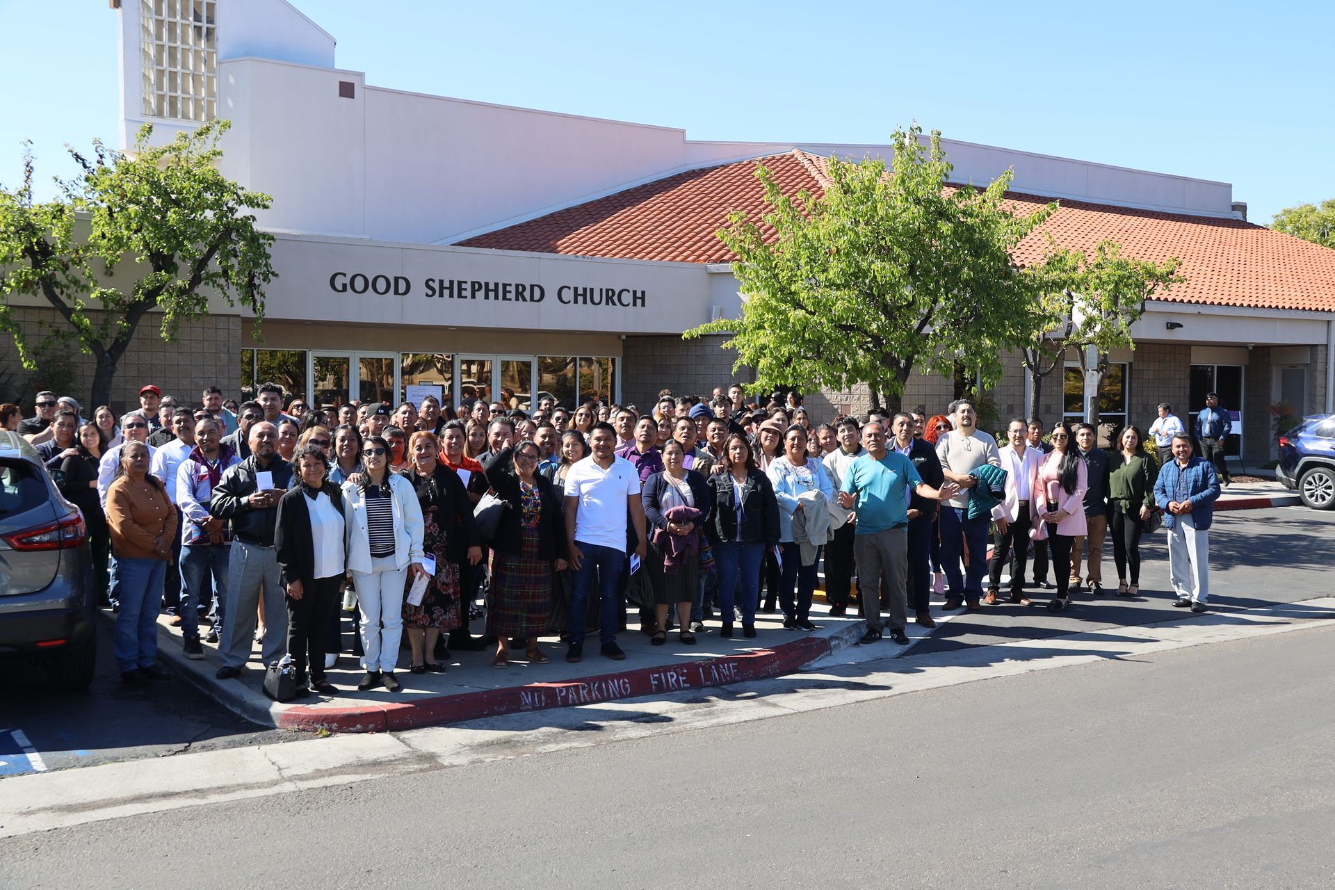 Crowd gathered outside Good Shepherd Church on a sunny day, standing in front of the building.