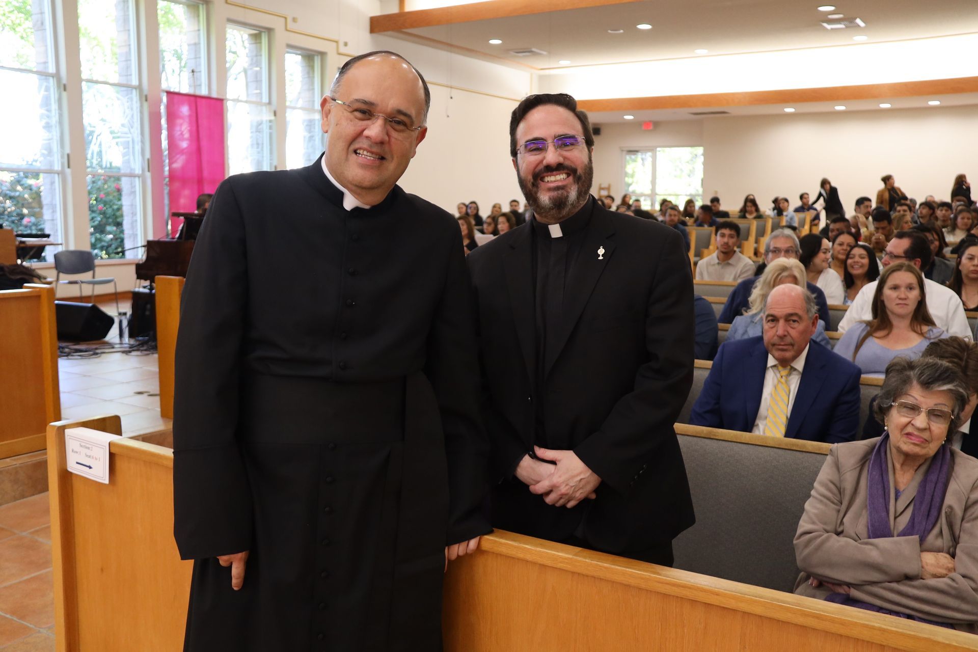 Two clergy members stand smiling in a church pew with a seated congregation behind them.